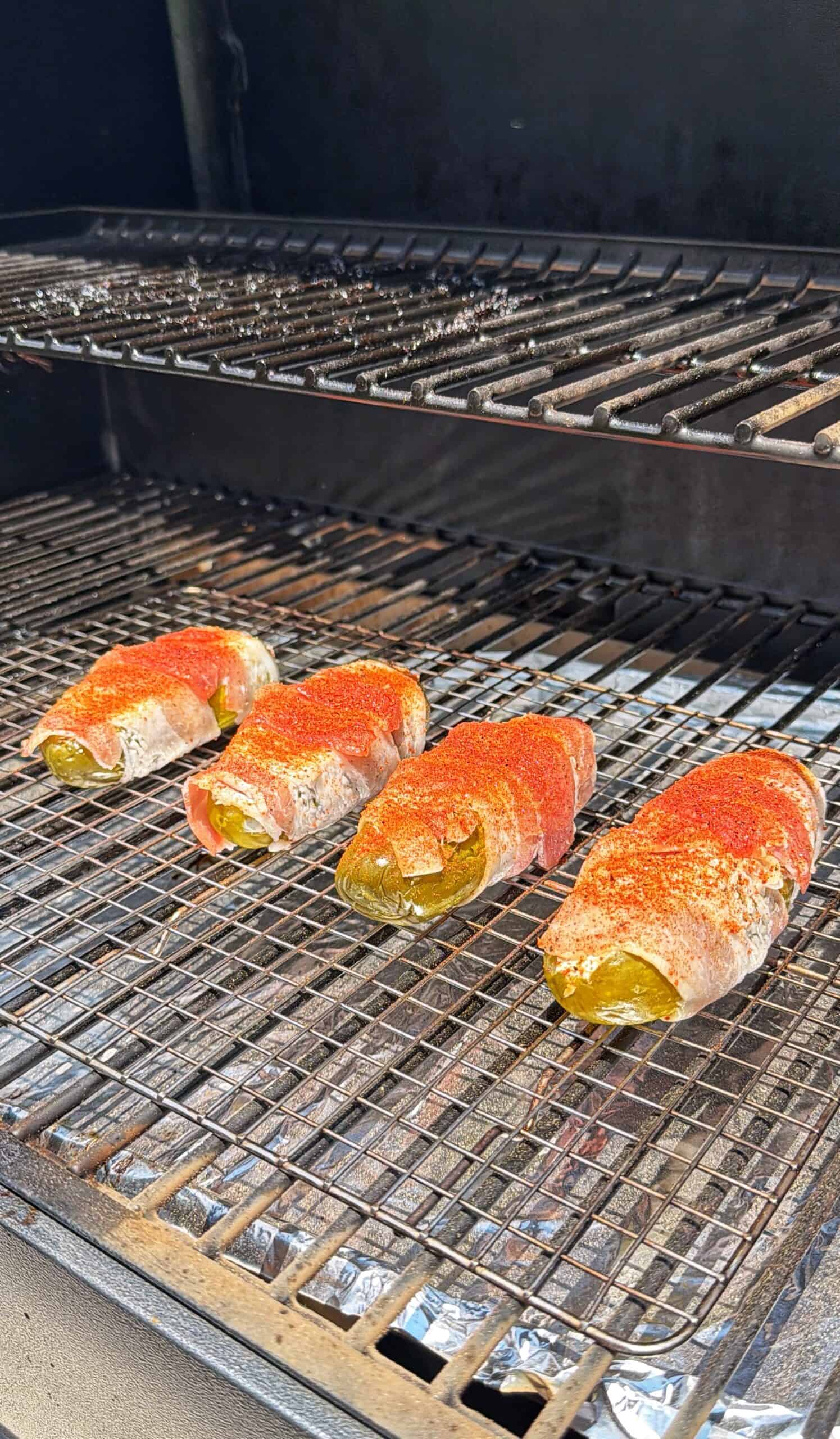 Four bacon-wrapped jalapeños, sprinkled with seasoning, are cooking on a wire rack inside a smoker or grill. Foil covers the lower rack.