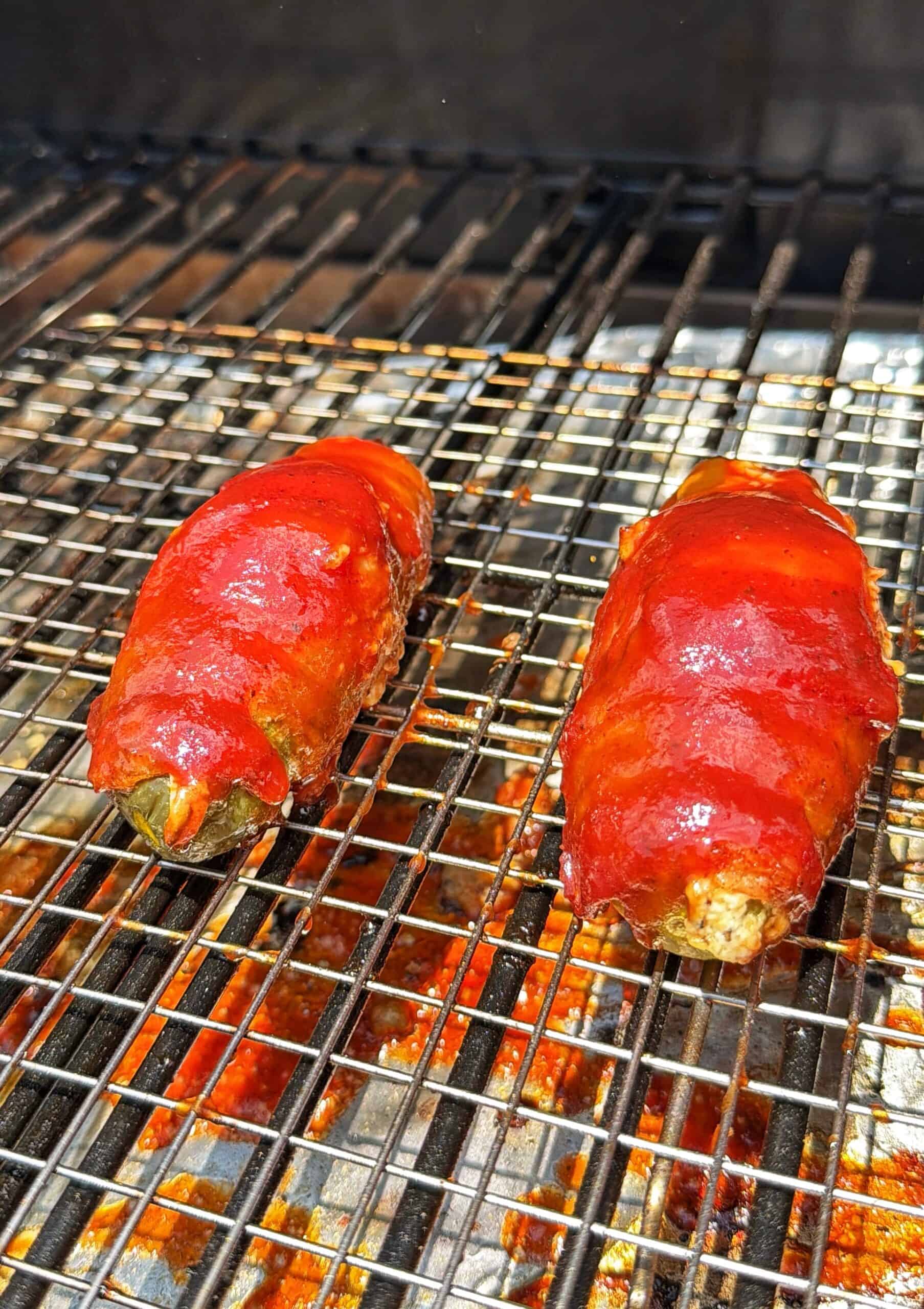 Two bacon-wrapped jalapeños stuffed with filling and coated in red sauce sit on a wire grill rack above hot coals.