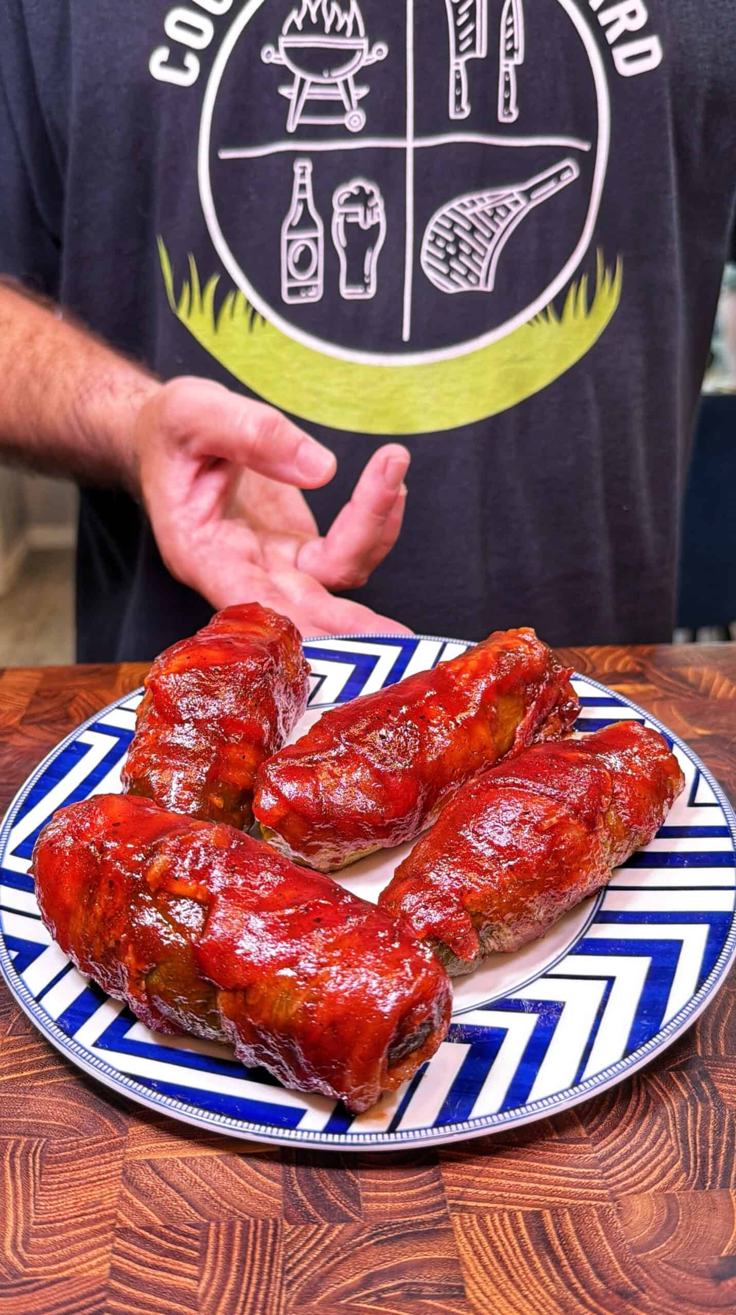 A person in a dark t-shirt presents a plate with four large, saucy barbecue ribs on a blue and white patterned plate, placed on a wooden surface.