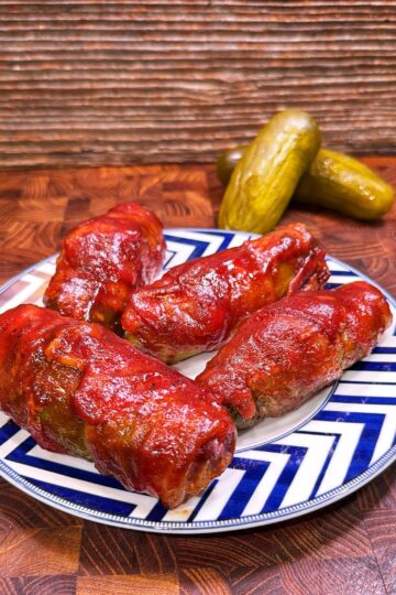 Four stuffed cabbage rolls topped with tomato sauce are arranged on a blue and white plate. In the background, three pickles rest on a wooden surface with a striped wooden backdrop.