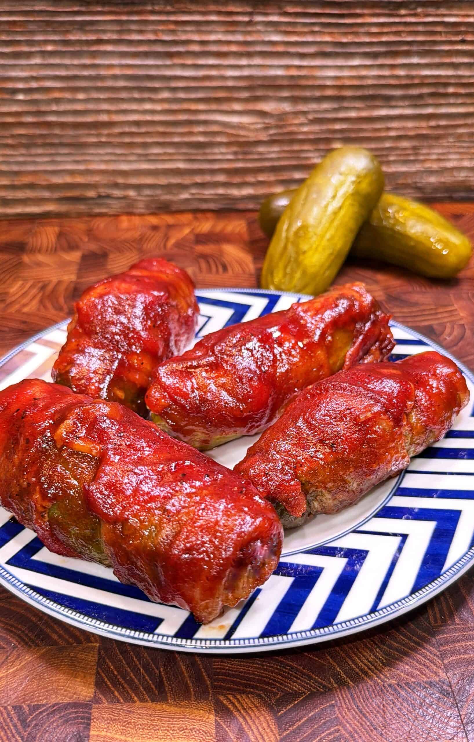 Four stuffed cabbage rolls topped with tomato sauce are arranged on a blue and white plate. In the background, three pickles rest on a wooden surface with a striped wooden backdrop.