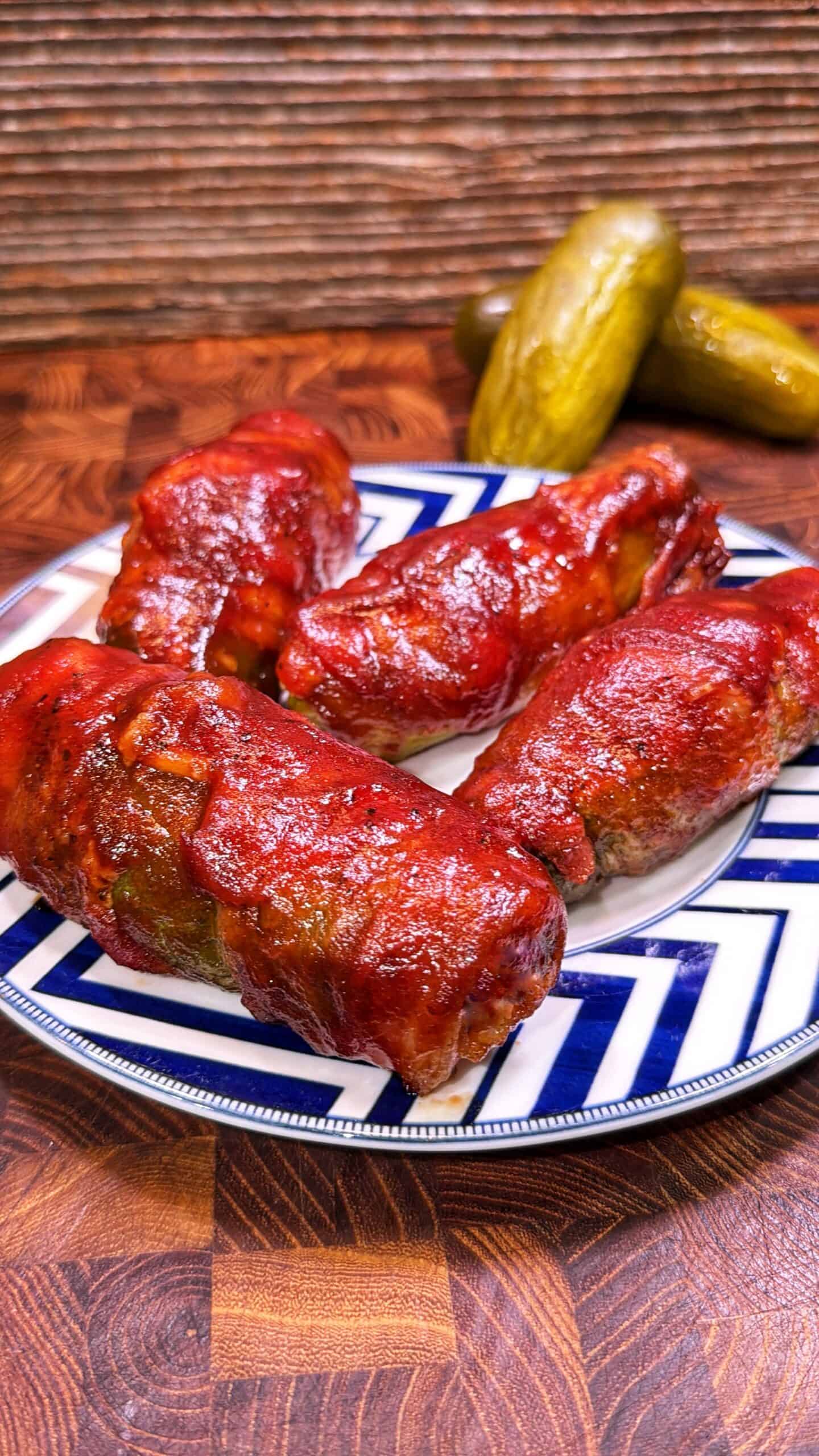 Four stuffed cabbage rolls covered in tomato sauce on a patterned plate, with two pickles in the background, set on a wooden surface.
