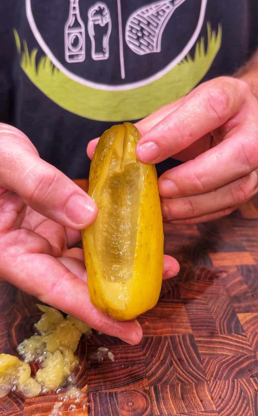 A person holds a hollowed-out pickle over a wooden surface, with pickle filling visible below. The person wears a shirt with food and drink graphics.