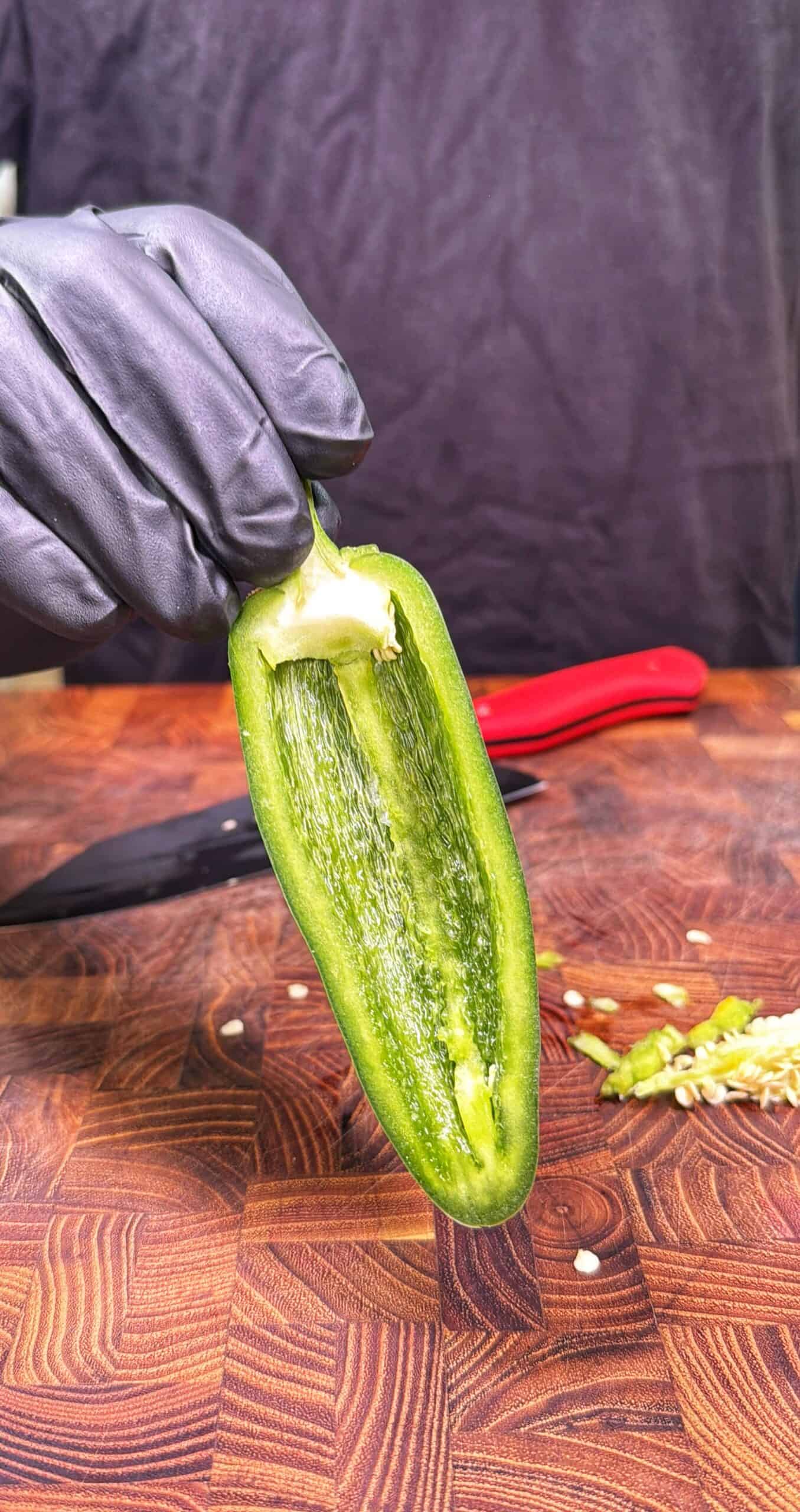 A gloved hand holds a halved jalapeño pepper over a wooden cutting board, with a black knife and sliced pepper pieces visible in the background.