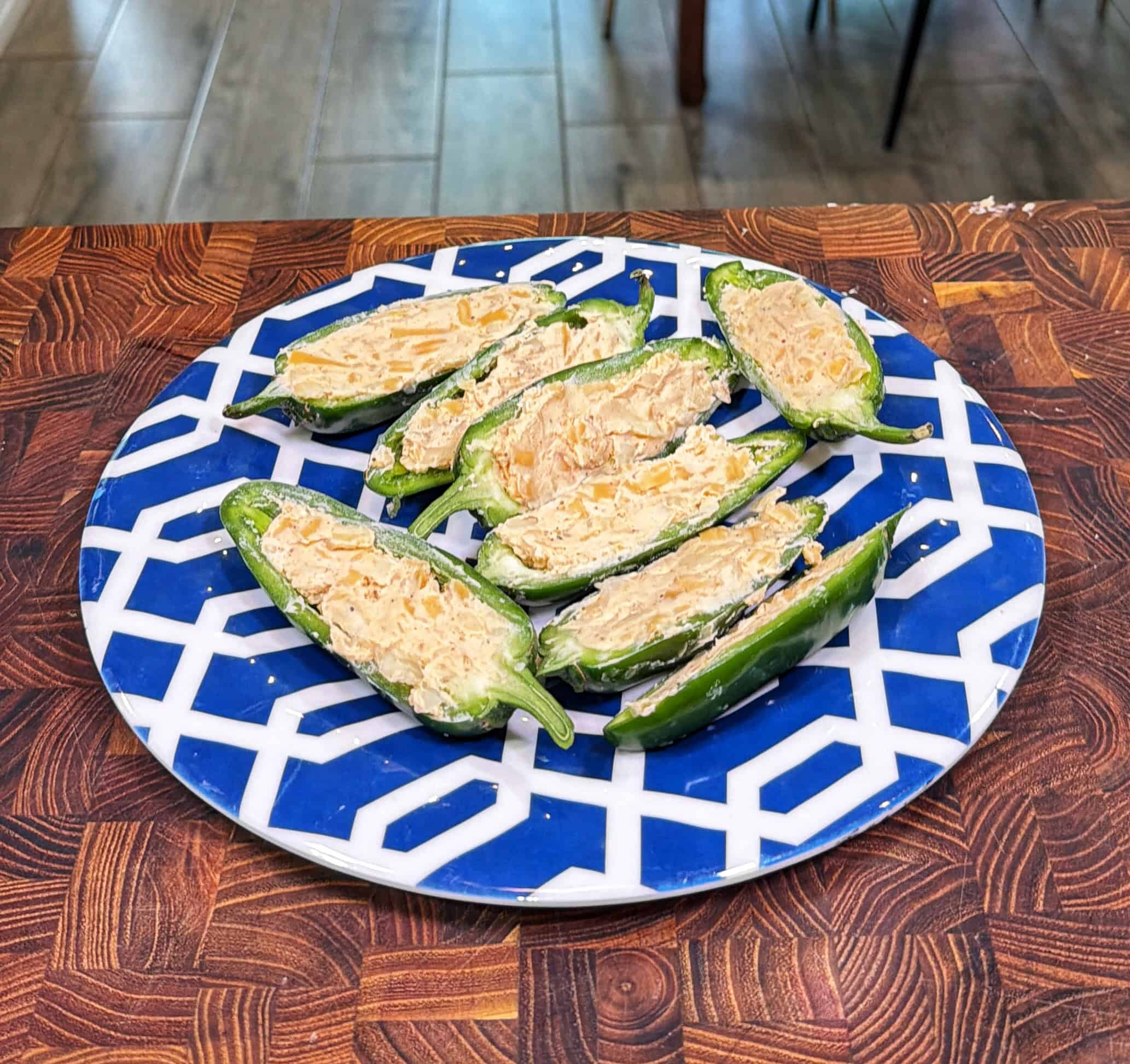 A plate of halved jalapeños filled with a creamy cheese mixture, arranged on a blue and white geometric-patterned plate, sits on a wooden surface.