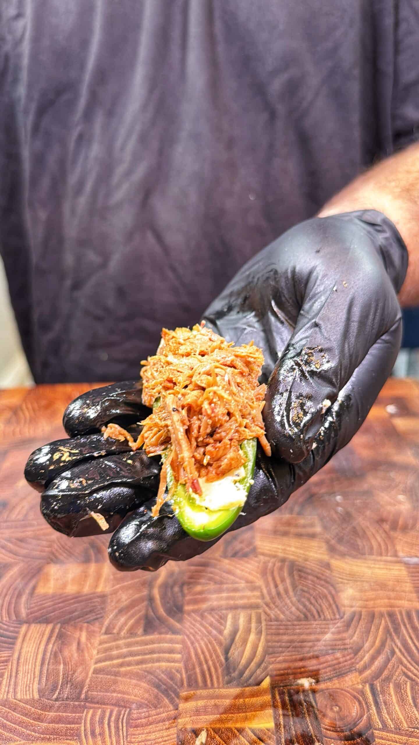 A person wearing a black glove holds a jalapeño half filled with shredded meat and cream cheese over a wooden cutting board.