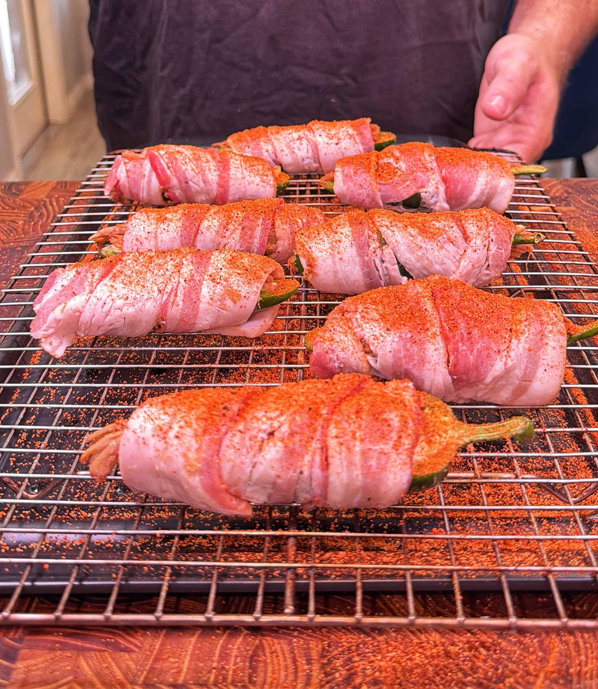 Eight bacon-wrapped jalapeños, dusted with seasoning, are arranged on a wire rack over a <a href=