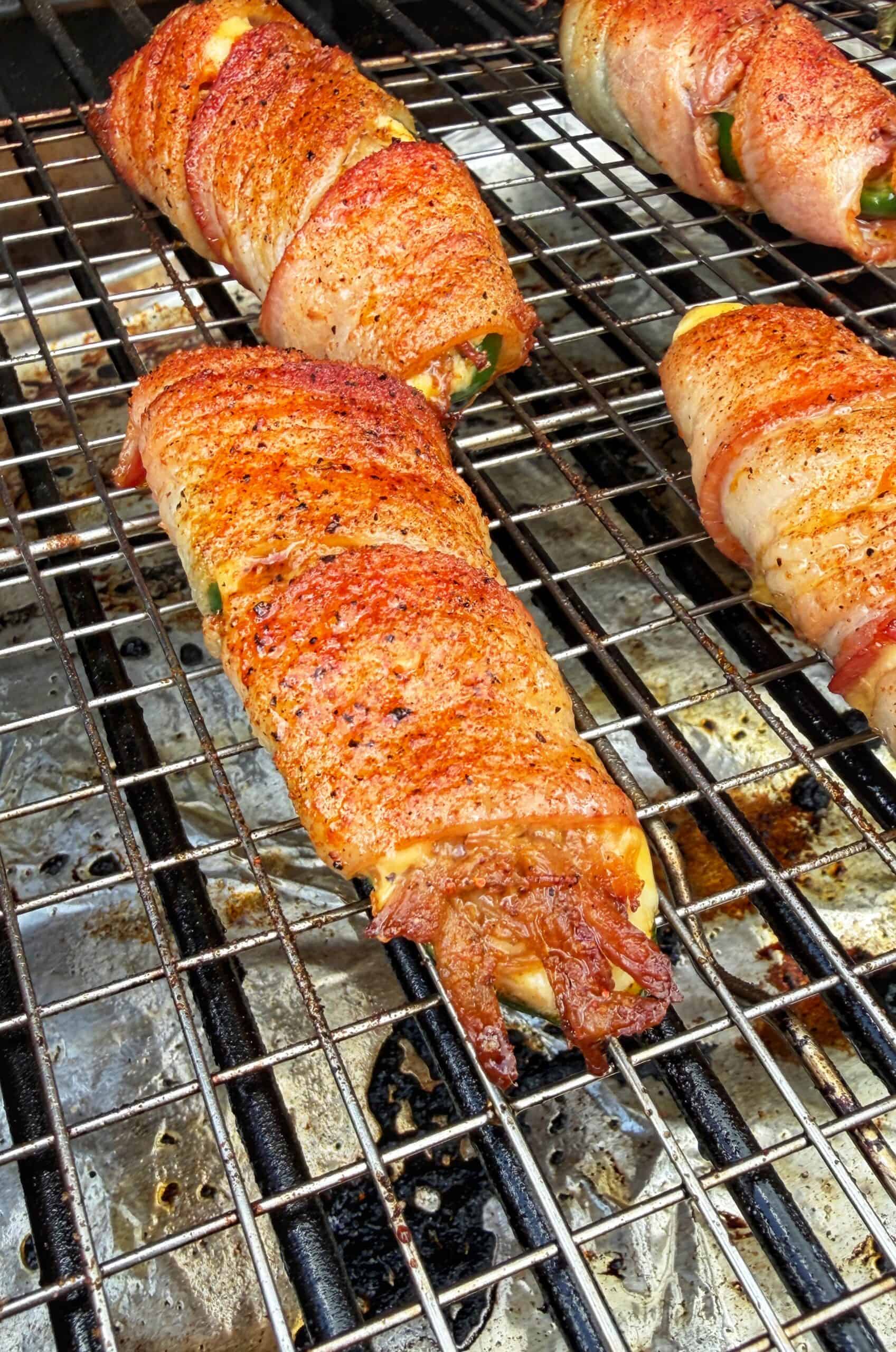 Bacon-wrapped, seasoned jalapeño poppers grilling on a metal rack, with melted cheese and crispy bacon edges visible.