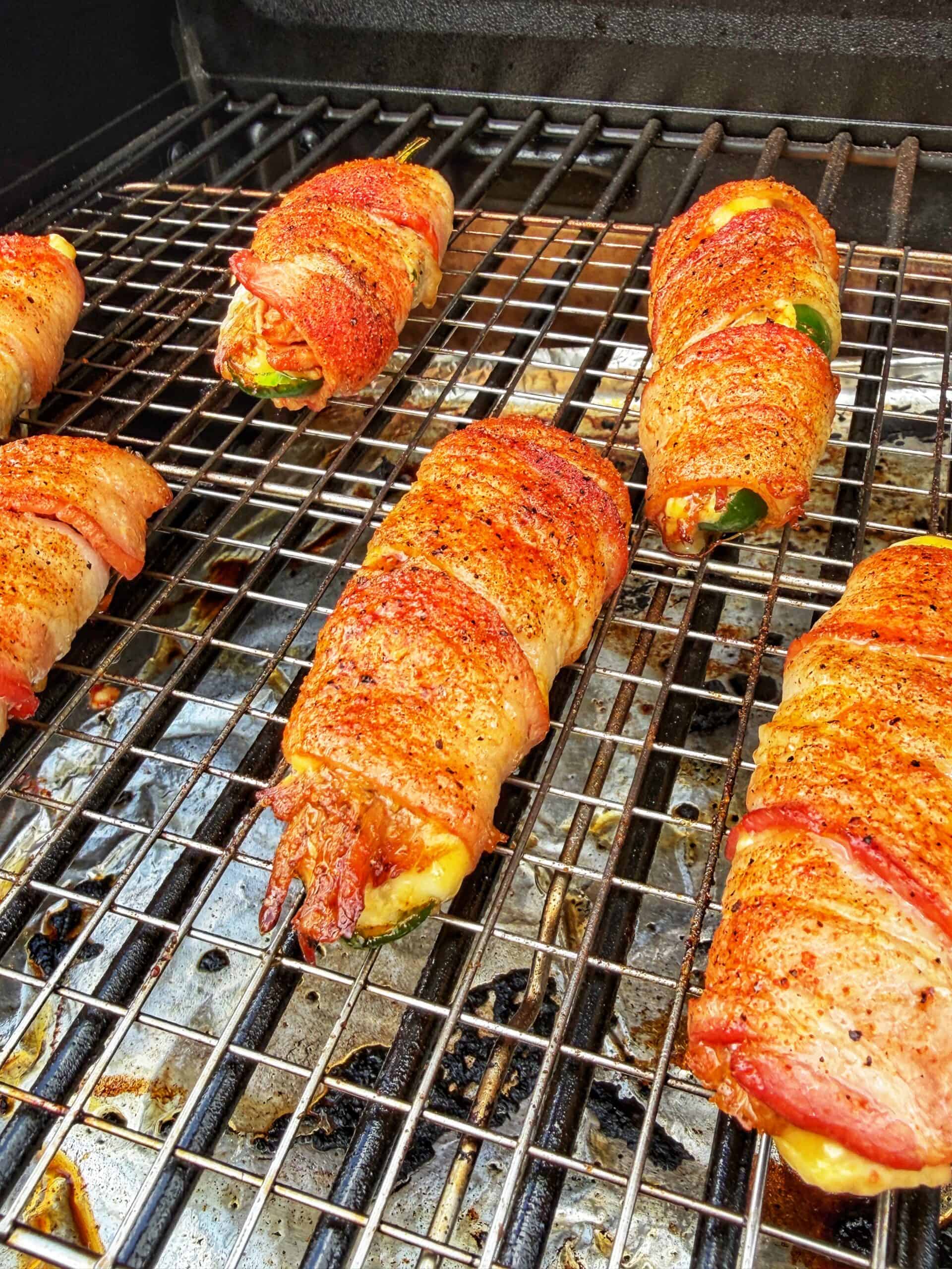 Six bacon-wrapped jalapeños stuffed with cheese and spices are grilling on a barbecue, showing crispy, golden-brown bacon and melted cheese peeking from the sides on a clean metal grill.