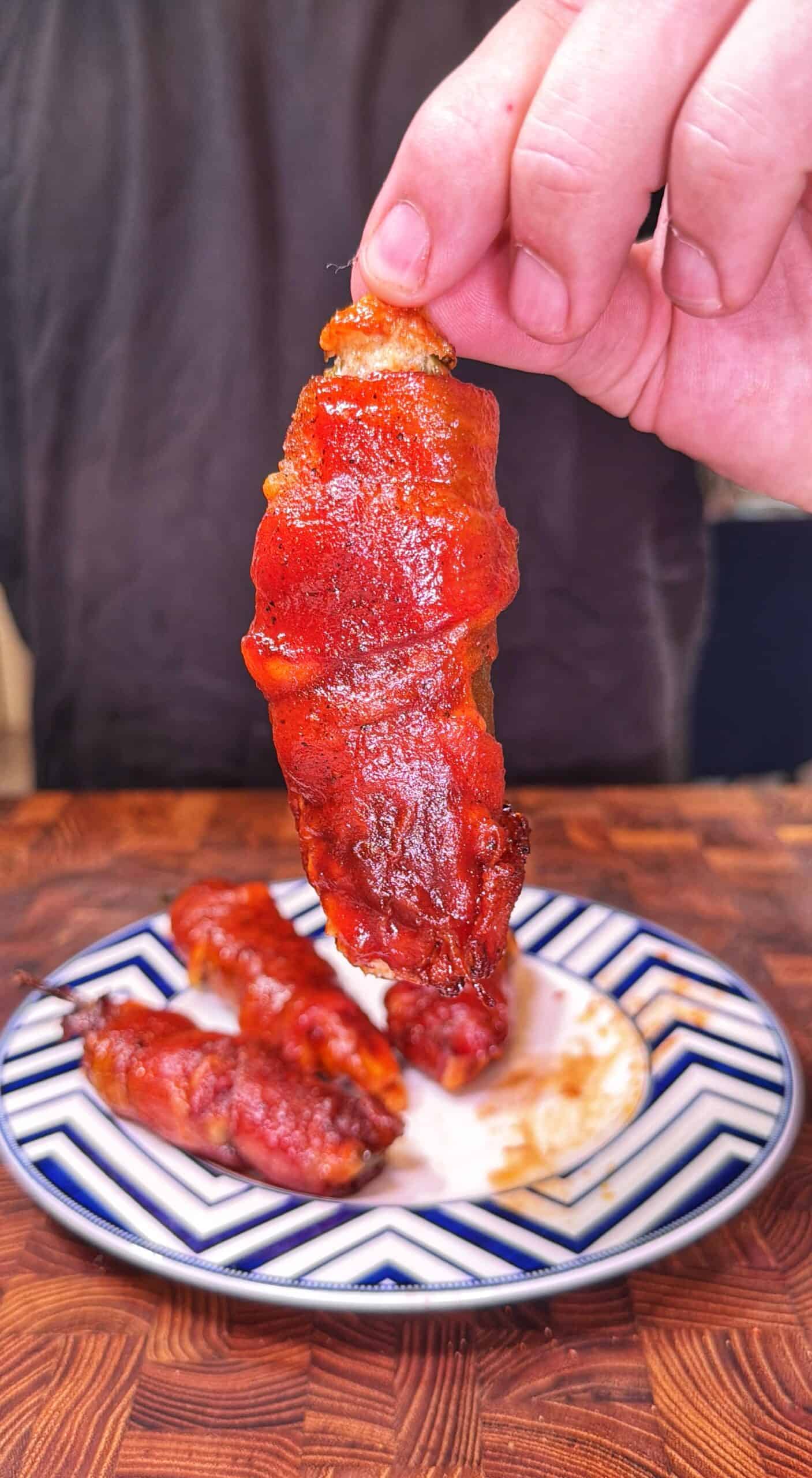 A hand holds up a saucy, cooked chicken wing over a plate with two more wings on a blue and white striped dish, all on a wooden table.
