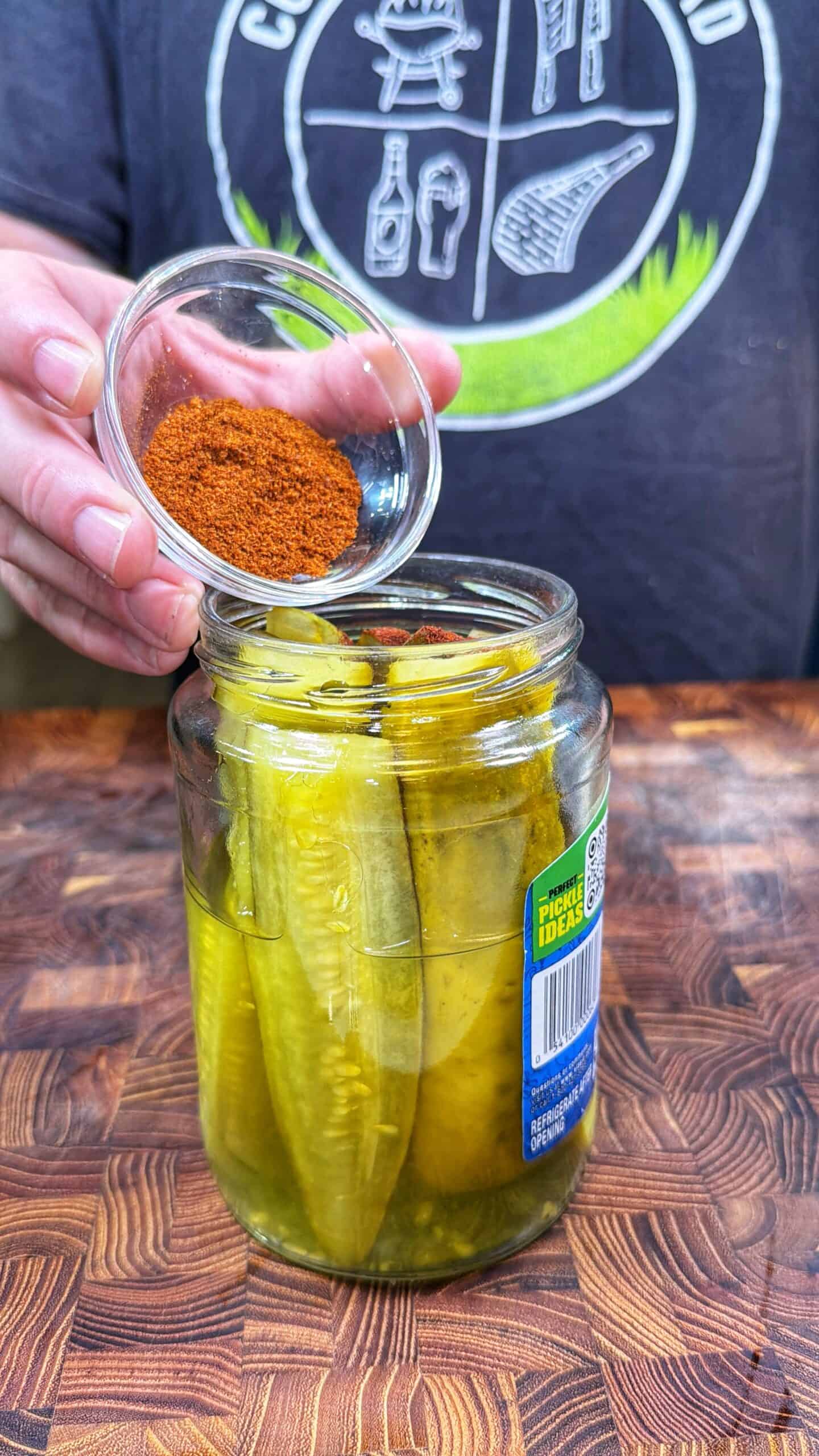 A person pours a small bowl of red seasoning into a jar of pickle spears on a wooden surface. The person is wearing a dark shirt with a logo on it.