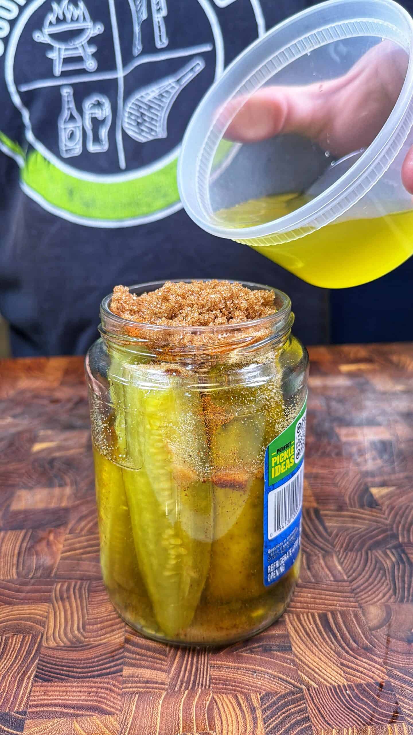 A hand pours yellow liquid into a jar of pickle spears topped with brown sugar, on a wooden surface. A person wearing a dark shirt with green and white graphics is in the background.