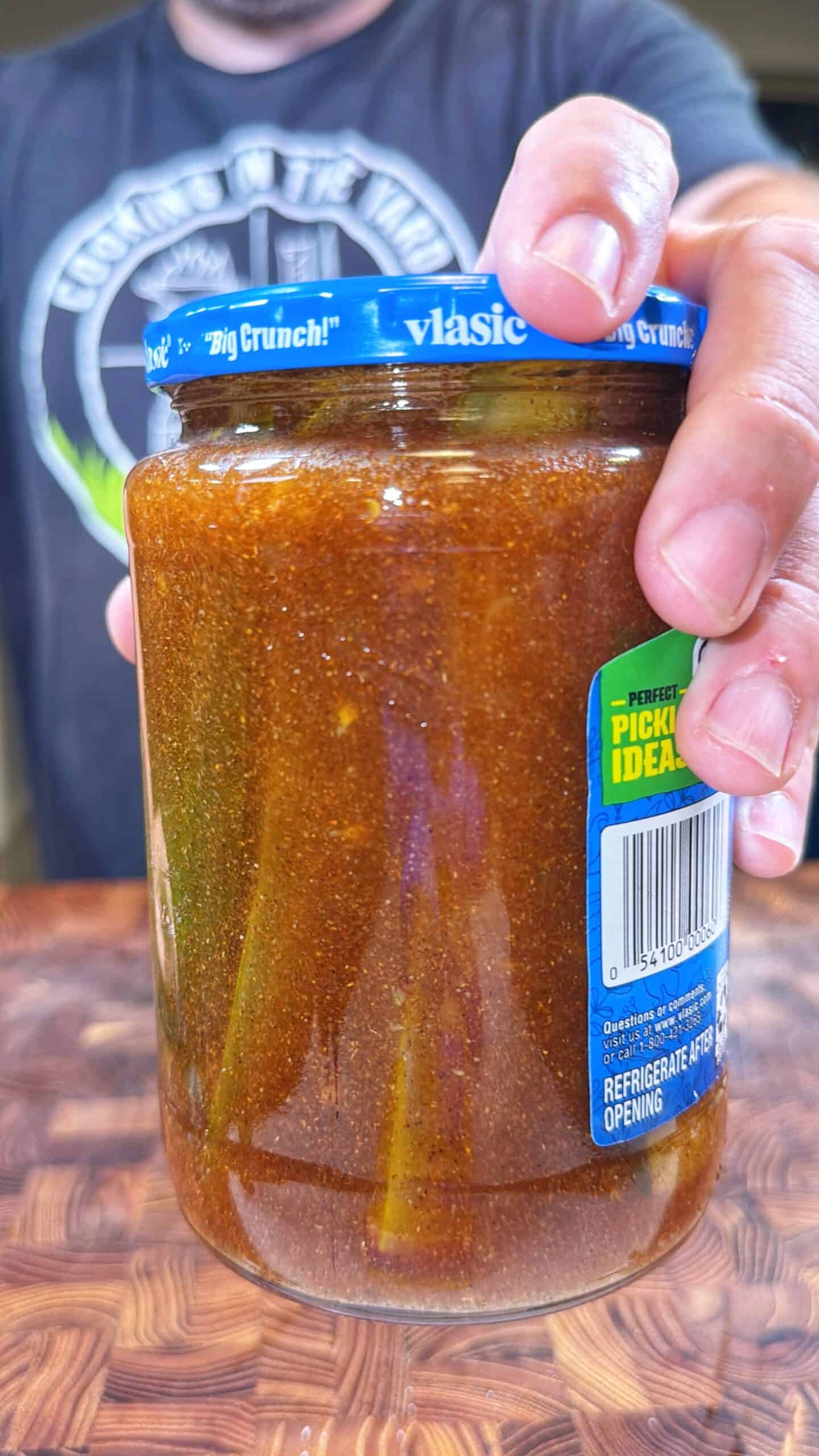 A person holds a Vlasic pickle jar filled with dark, seasoned liquid and visible pickle spears inside, over a wooden surface.
