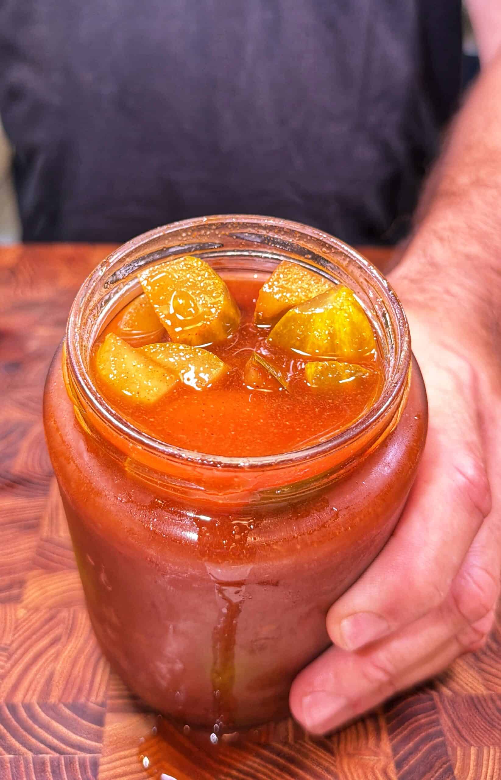 A hand holds a jar filled with reddish liquid and chunks of green pickles, sitting on a wooden surface.