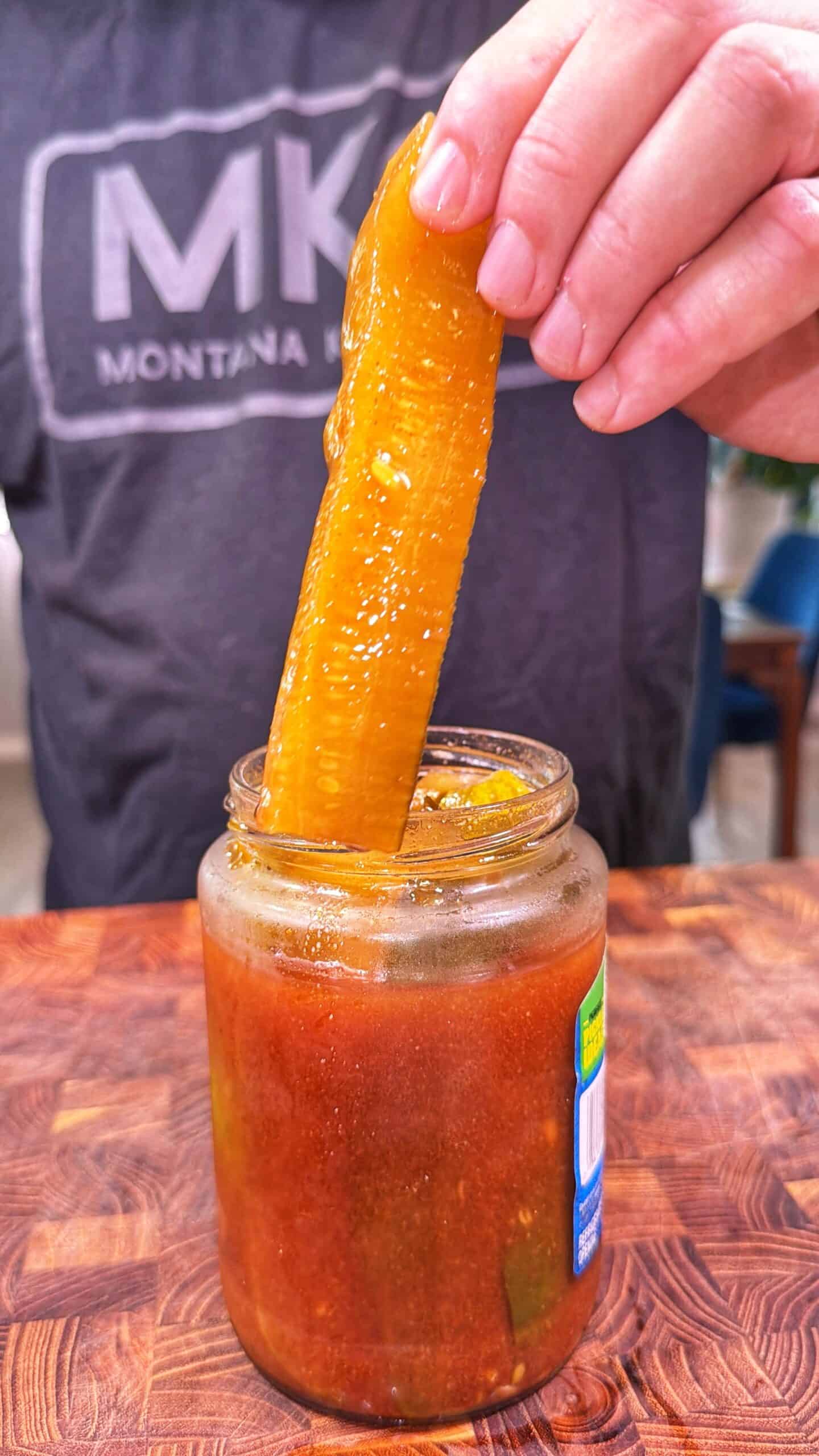 A hand lifts a large pickle from a jar filled with reddish liquid on a wooden surface. The person is wearing a gray shirt with MK MONTANA printed on it.