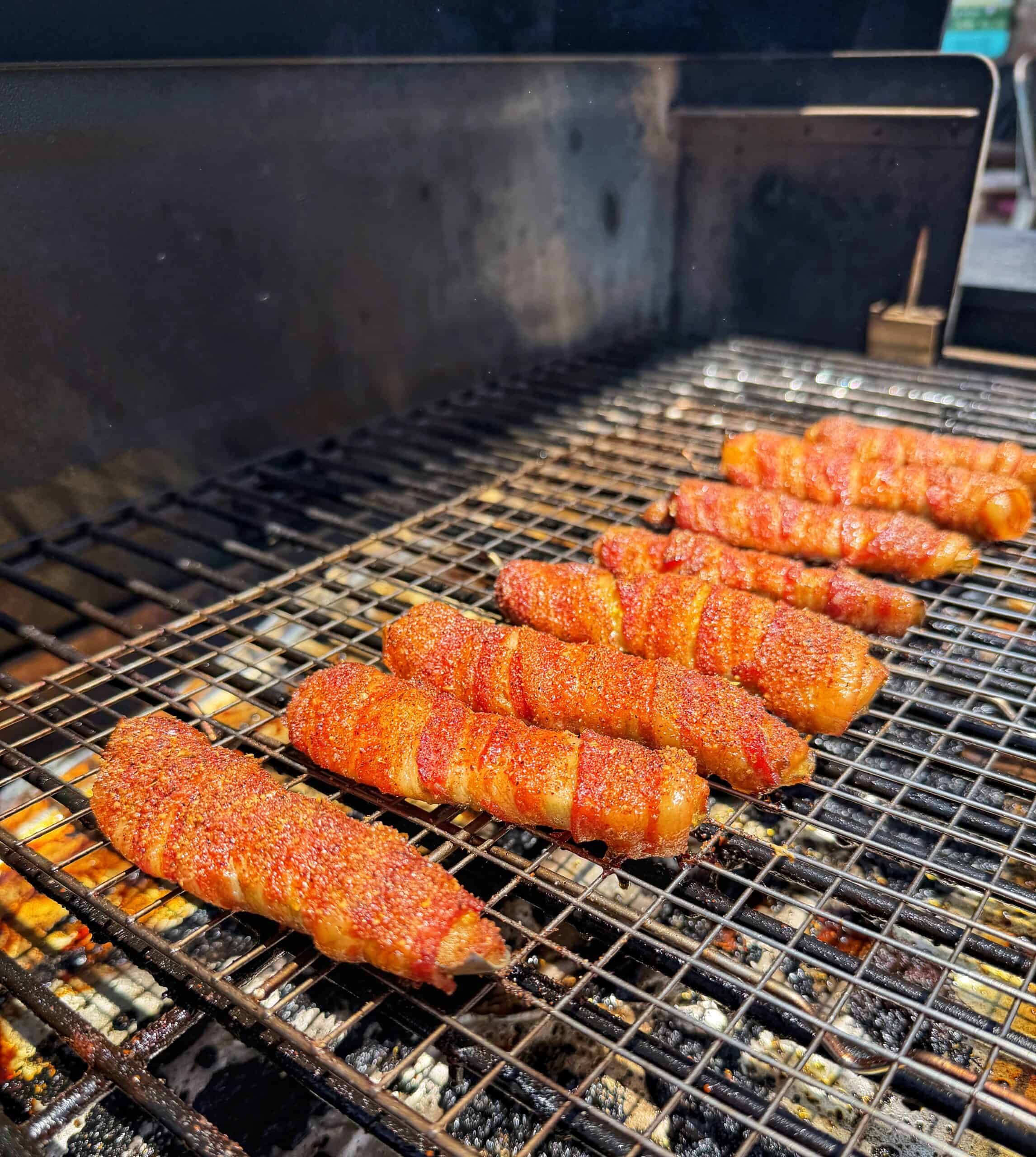 Several bacon-wrapped pieces of meat, sprinkled with seasoning, are grilling on a metal rack inside a barbecue smoker. The food is evenly spaced, and the grill marks are visible beneath.