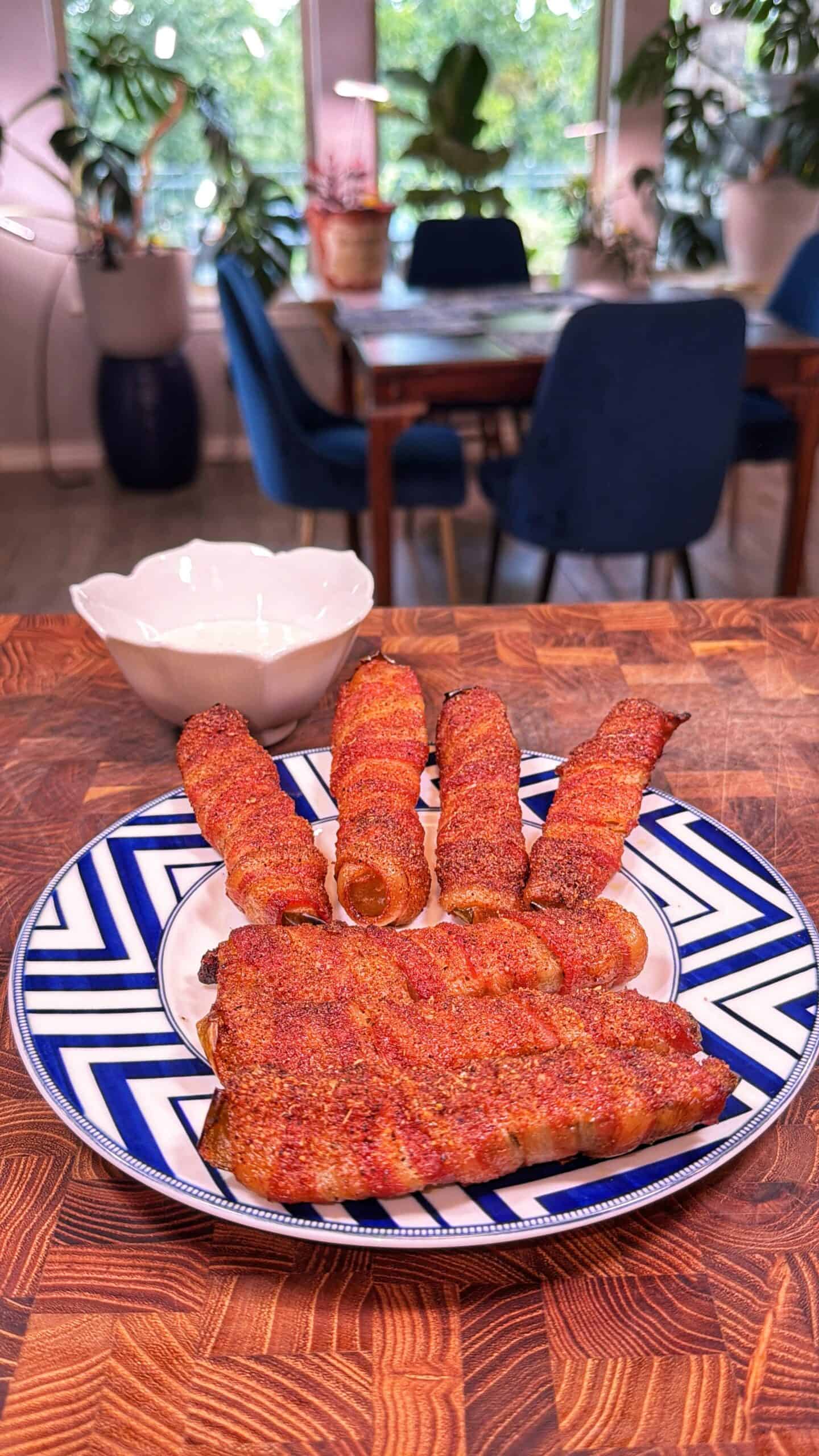 A plate with bacon-wrapped sausages and a slab of seasoned meat sits on a patterned blue and white plate. A white bowl is nearby, and a dining area with blue chairs and plants is in the background.
