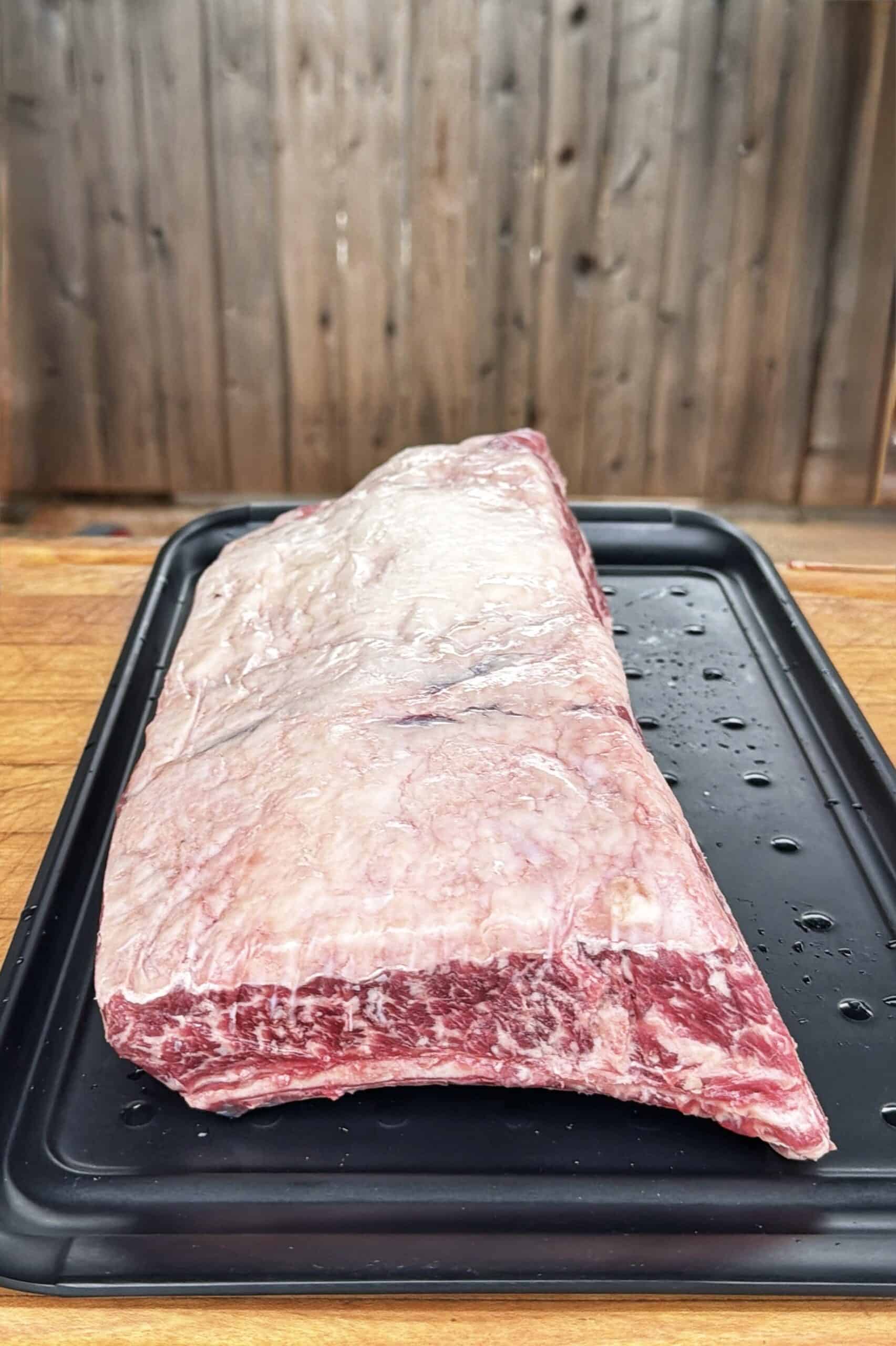 A large, raw, marbled beef brisket sits on a black tray atop a wooden surface, with a wooden fence in the blurred background.