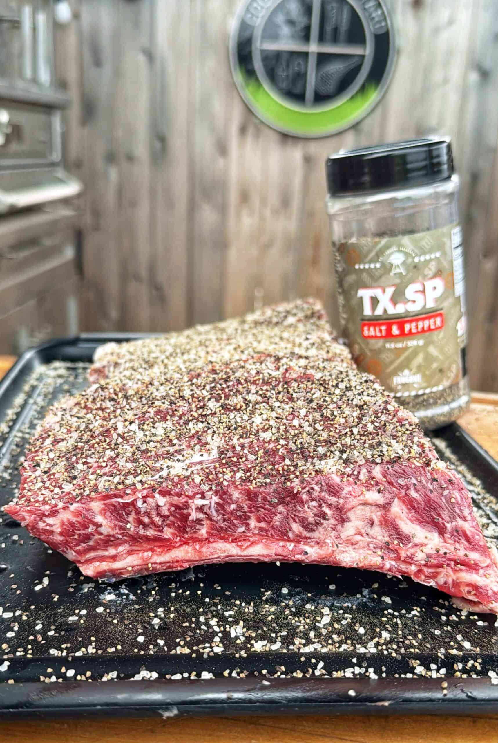 Close-up of raw beef brisket covered in coarse salt and pepper seasoning on a tray, with a container of TX.SP Salt & Pepper seasoning visible in the background.