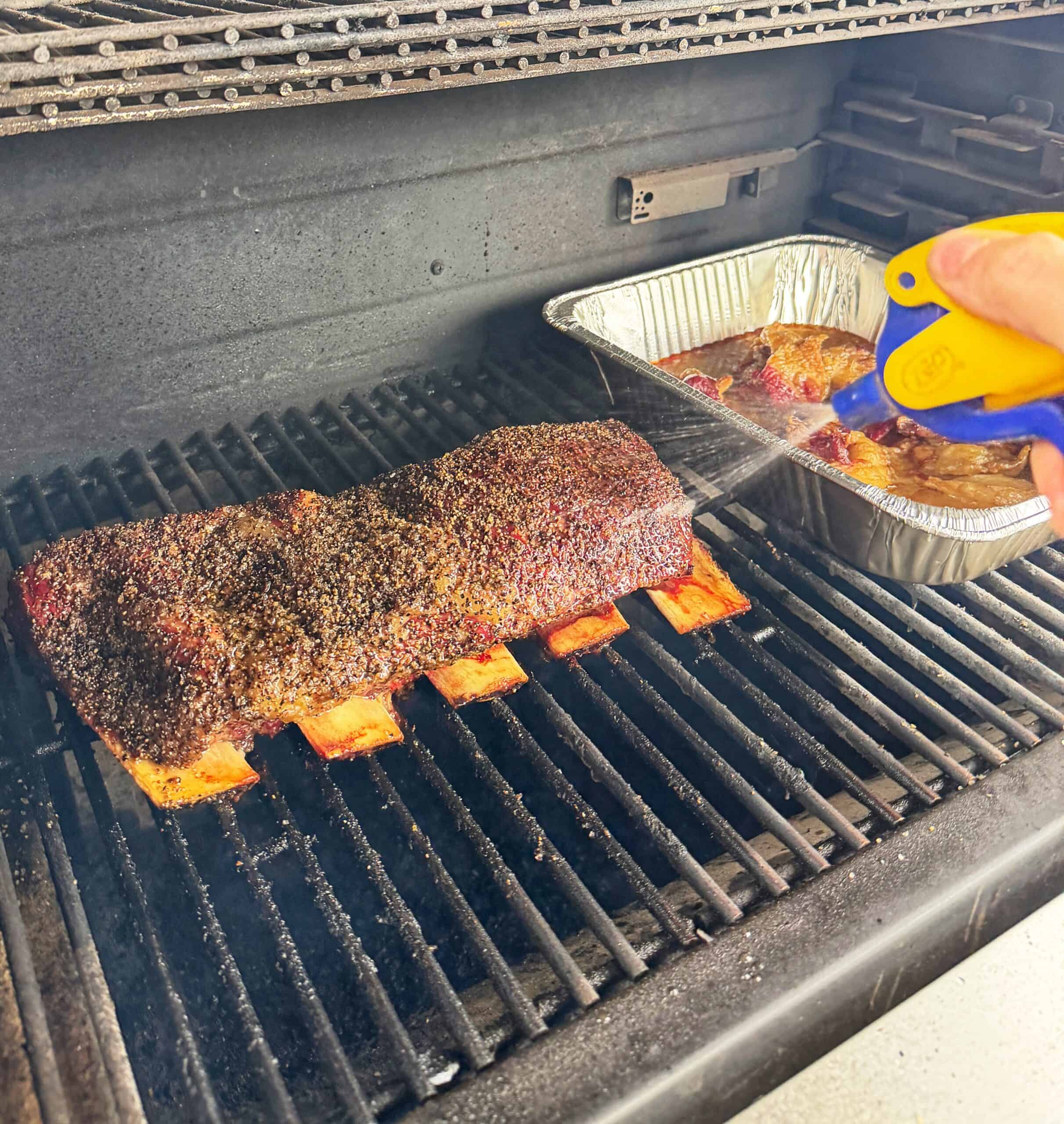 A hand sprays liquid onto beef ribs cooking in a smoker. The ribs are seasoned, and a foil pan with meat sits nearby. Smoke is visible rising inside the smoker.