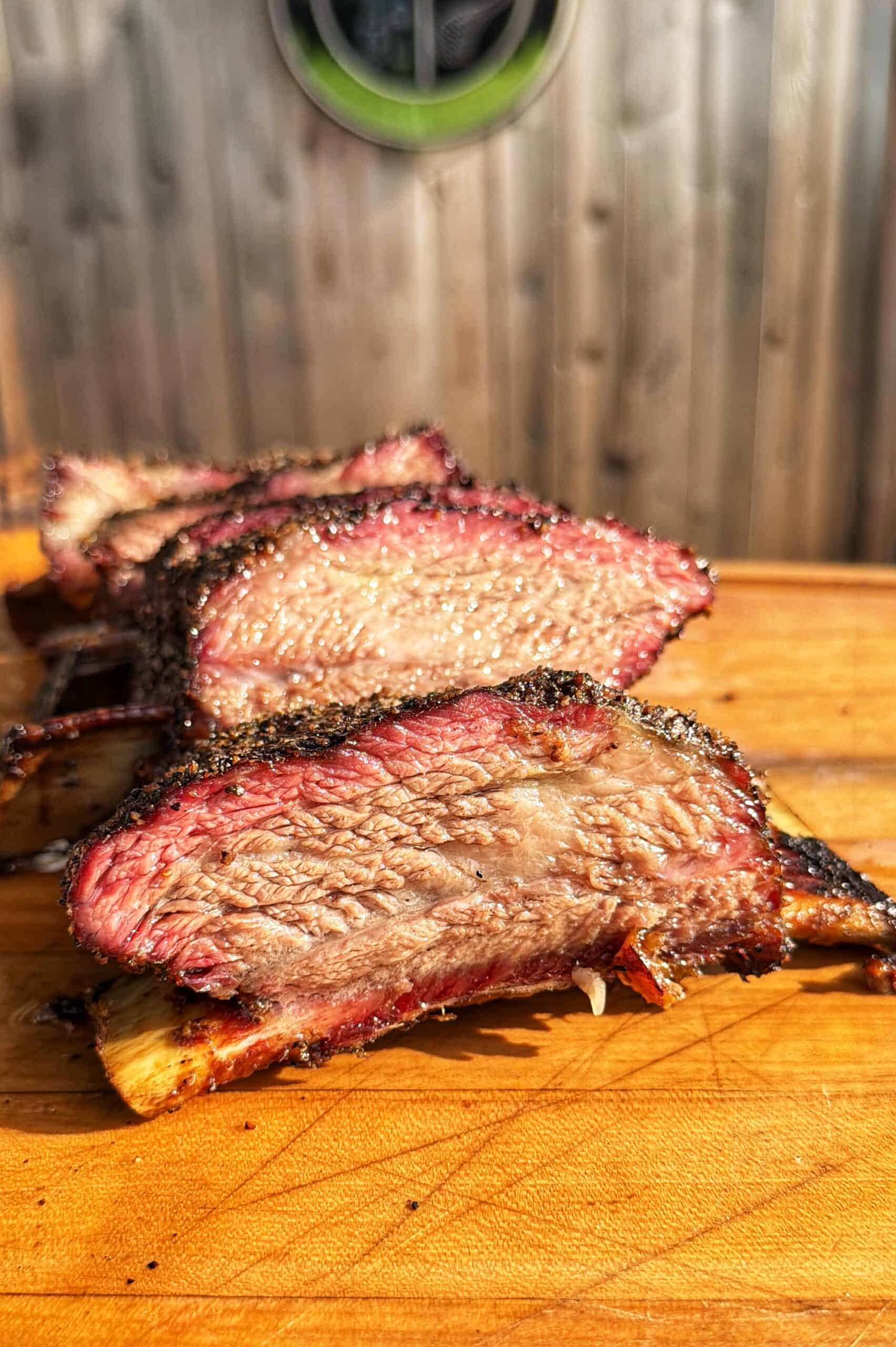 Three thick slices of smoked beef brisket with a visible smoke ring sit on a wooden cutting board in front of a wooden wall. The meat looks juicy and seasoned, with a crispy outer bark.