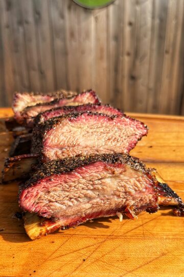 Four thick, juicy slices of seasoned smoked beef ribs are arranged on a wooden cutting board, with a sunlit wooden fence blurred in the background.