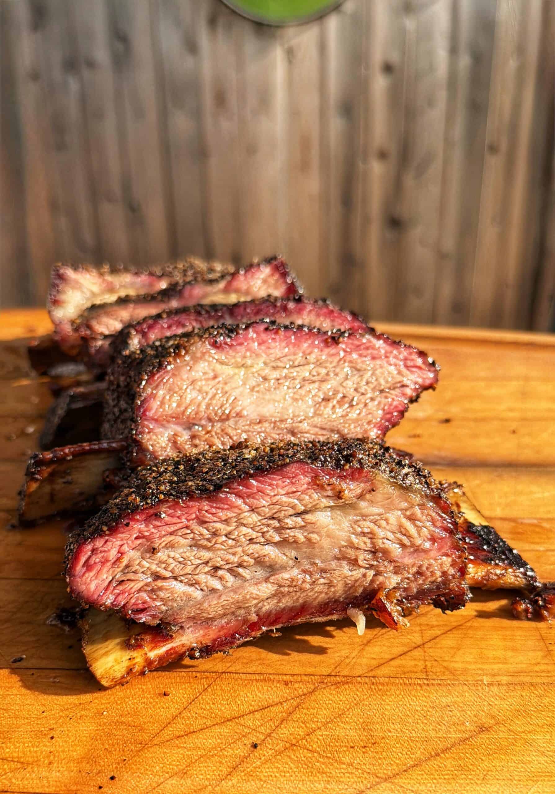 Four thick, juicy slices of seasoned smoked beef ribs are arranged on a wooden cutting board, with a sunlit wooden fence blurred in the background.