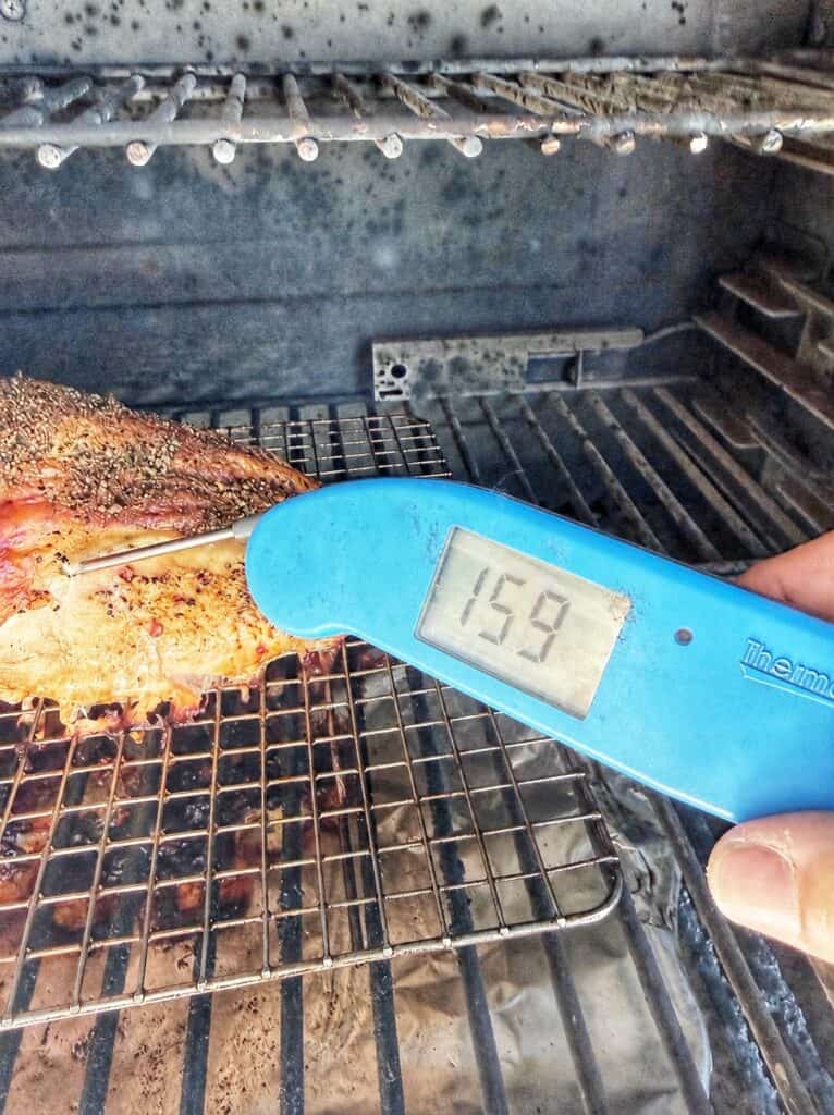 A digital meat thermometer reading 159°F in seasoned frog-cut chicken on a wire rack inside a grill.