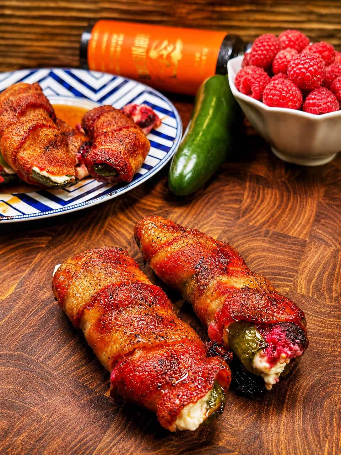 Two bacon-wrapped stuffed jalapeños on a wooden surface, with more on a plate, a bowl of raspberries, a whole jalapeño, and an orange bottle in the background.