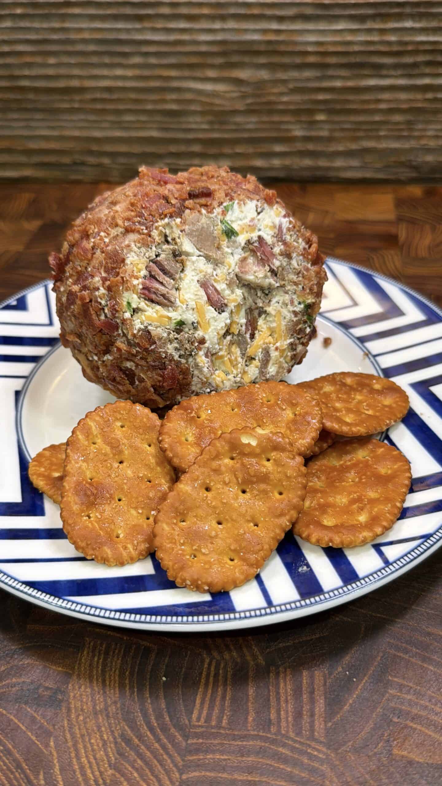 A round cheese ball coated with chopped pecans sits on a plate with several crispy, seasoned crackers. The plate features a blue and white geometric pattern.
