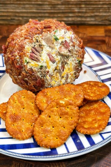A cheese ball coated with bacon and herbs sits on a patterned plate next to several round, golden crackers. The plate rests on a wooden table with a rustic background.