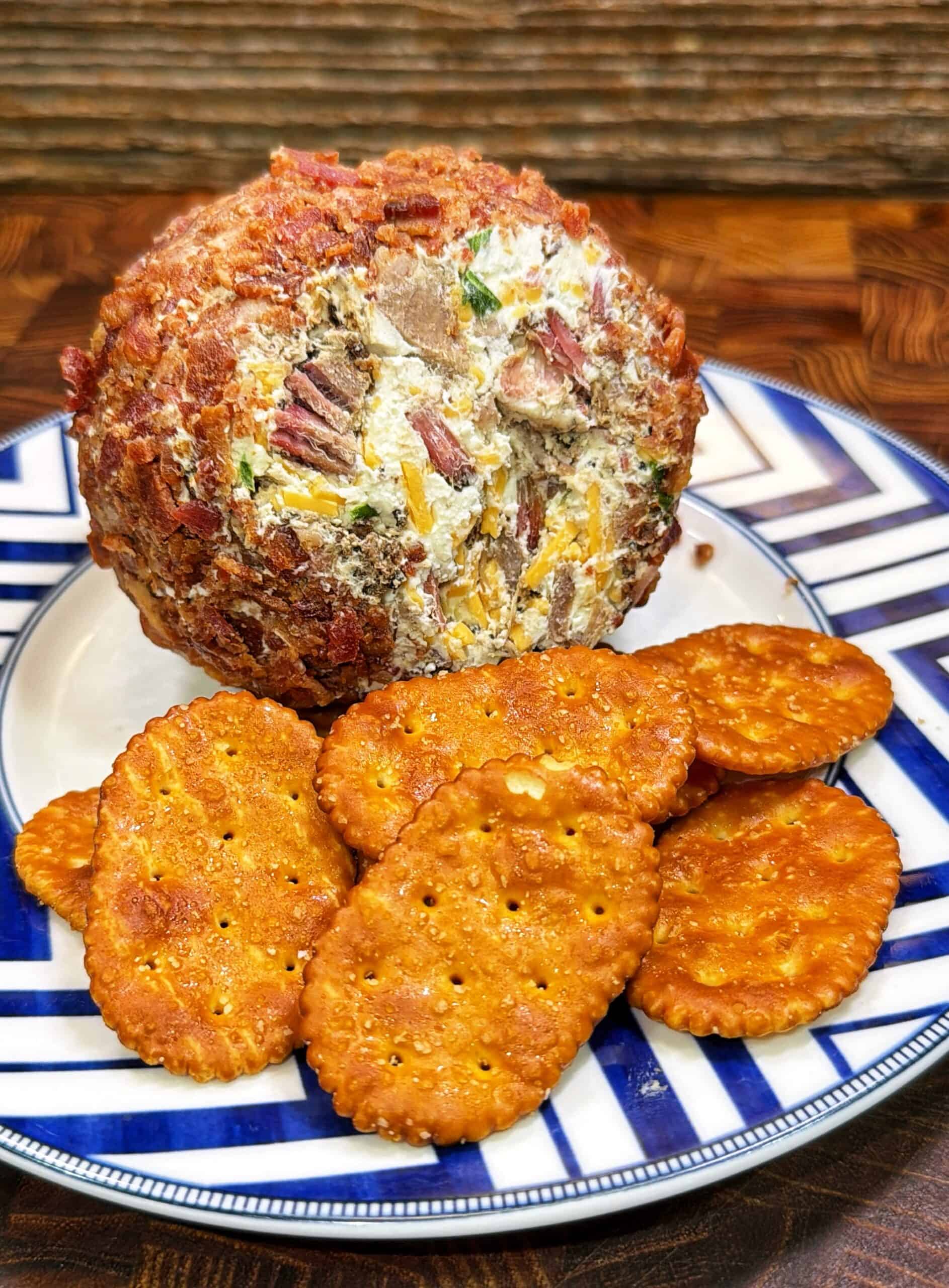 A cheese ball coated with bacon and herbs sits on a patterned plate next to several round, golden crackers. The plate rests on a wooden table with a rustic background.