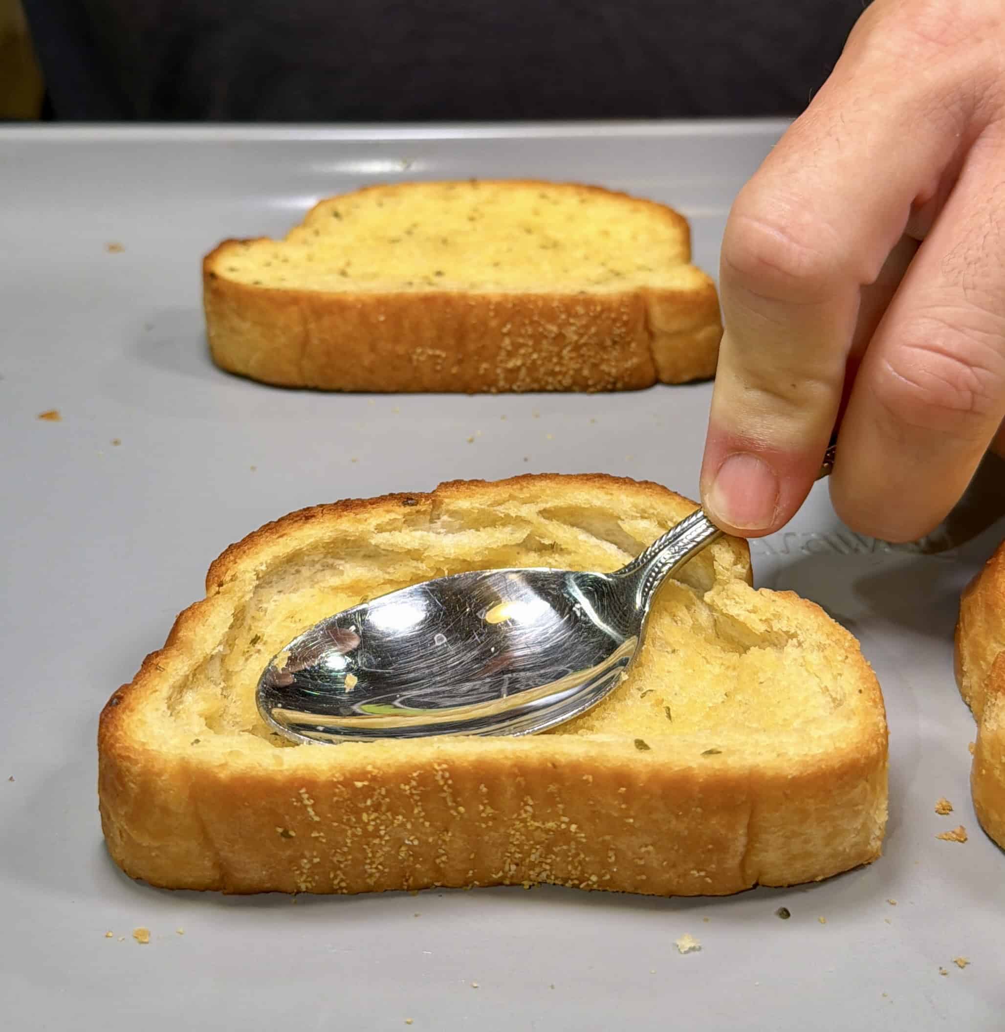 A hand presses a spoon into a slice of toasted bread on a gray tray, creating an indentation. Other slices of toast are in the background.