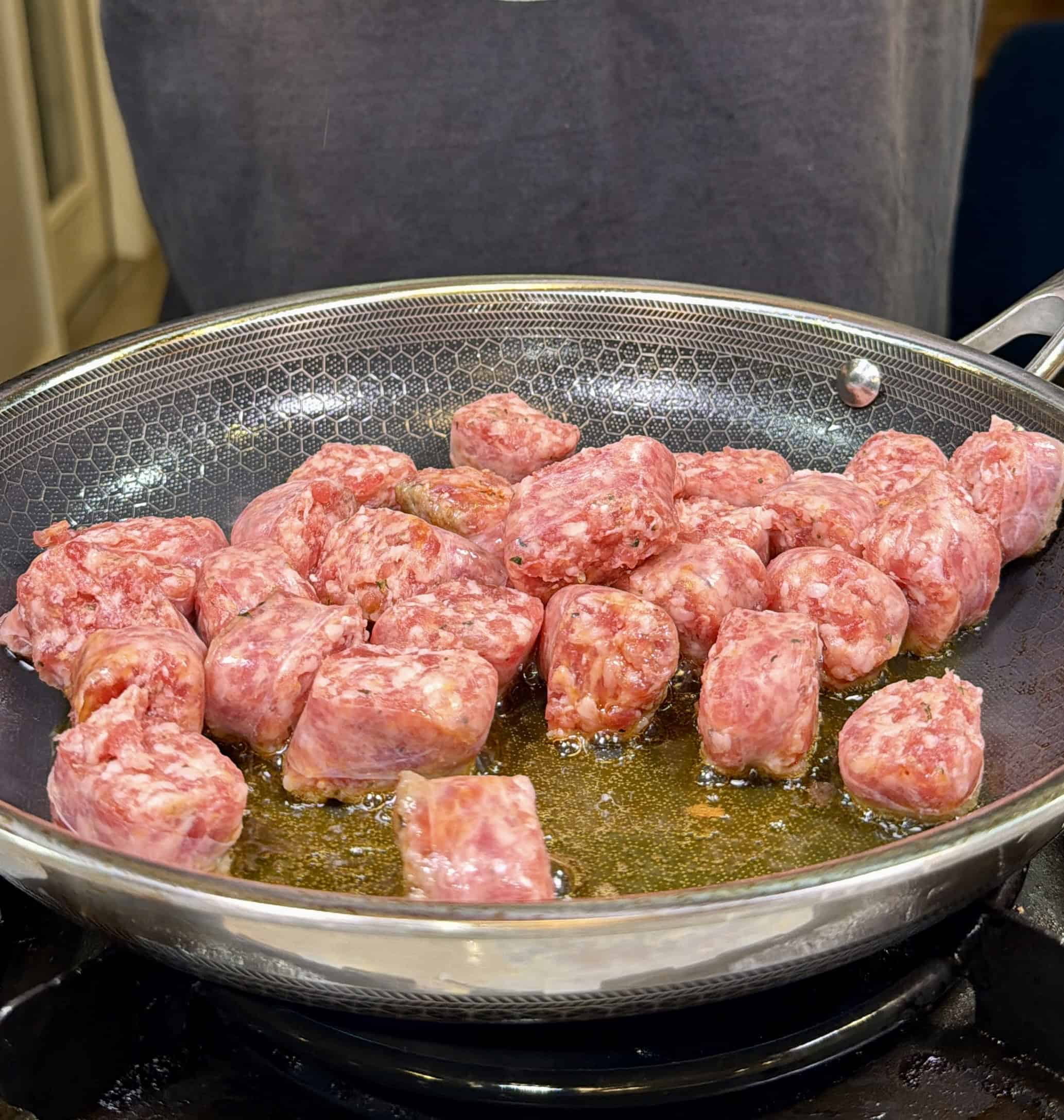Chunks of raw sausage are sizzling in oil in a stainless steel frying pan on a stovetop, with someone standing behind the pan.