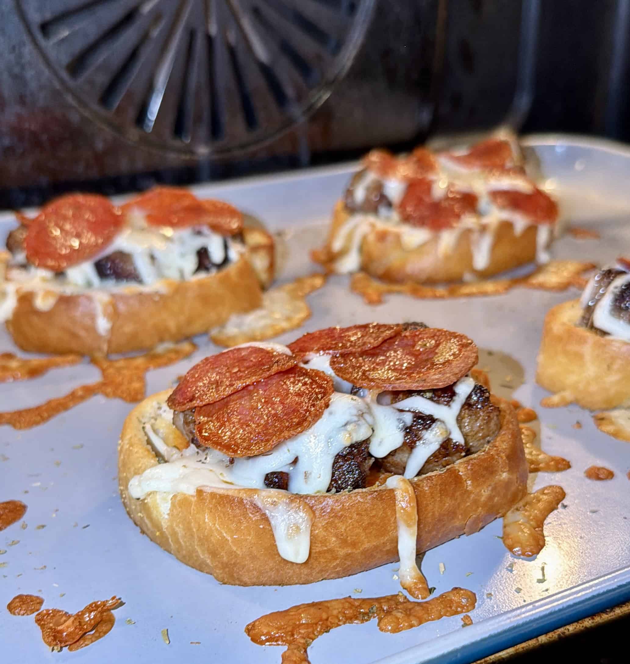 Slices of bread topped with melted cheese, pepperoni, and meat sit on a baking tray, fresh out of the oven. Cheese is melted and slightly browned, with some drippings on the tray.