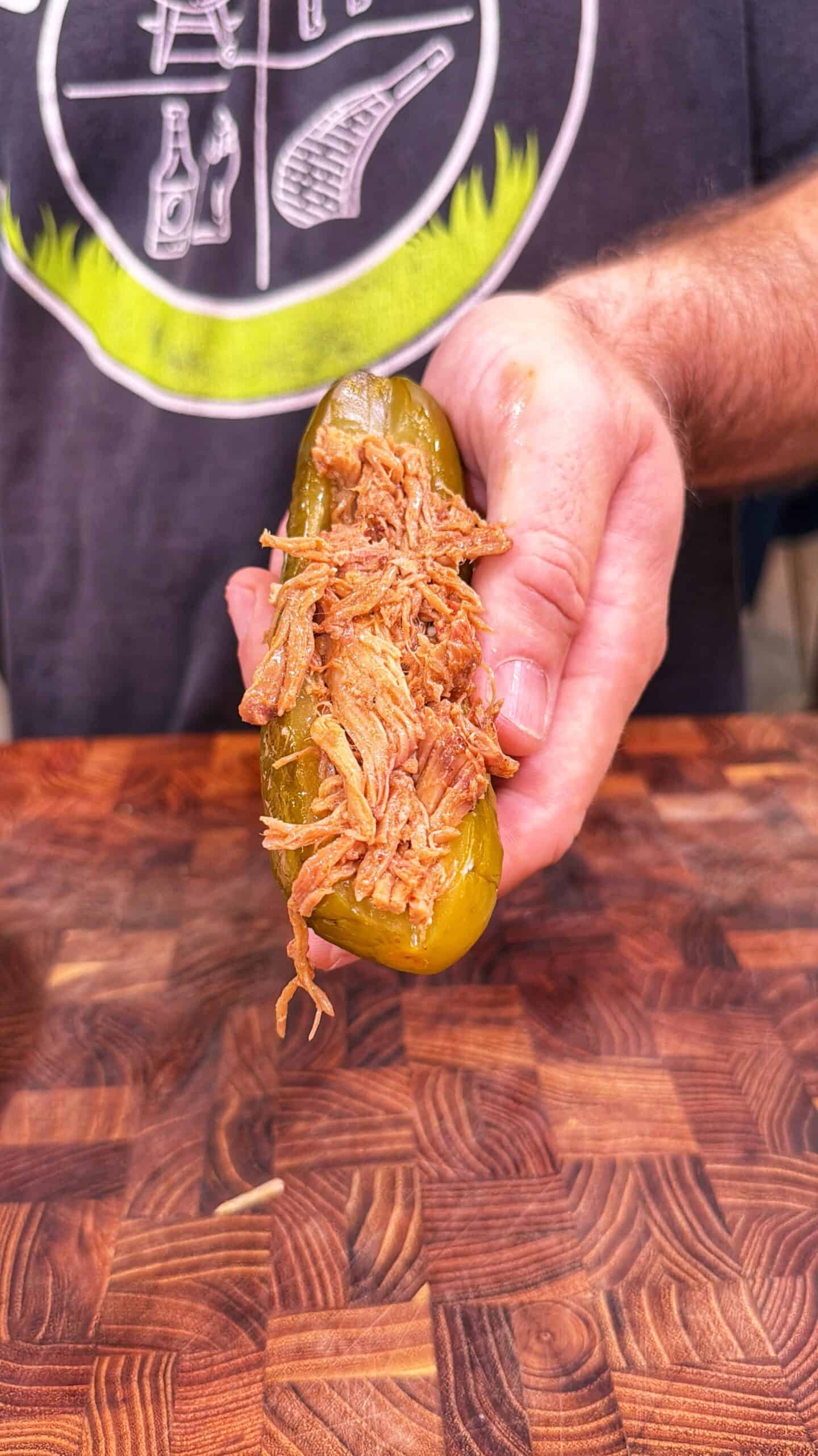 A person holds a pickle sliced open and stuffed with shredded meat over a wooden surface. The person wears a shirt with a barbecue-themed graphic.