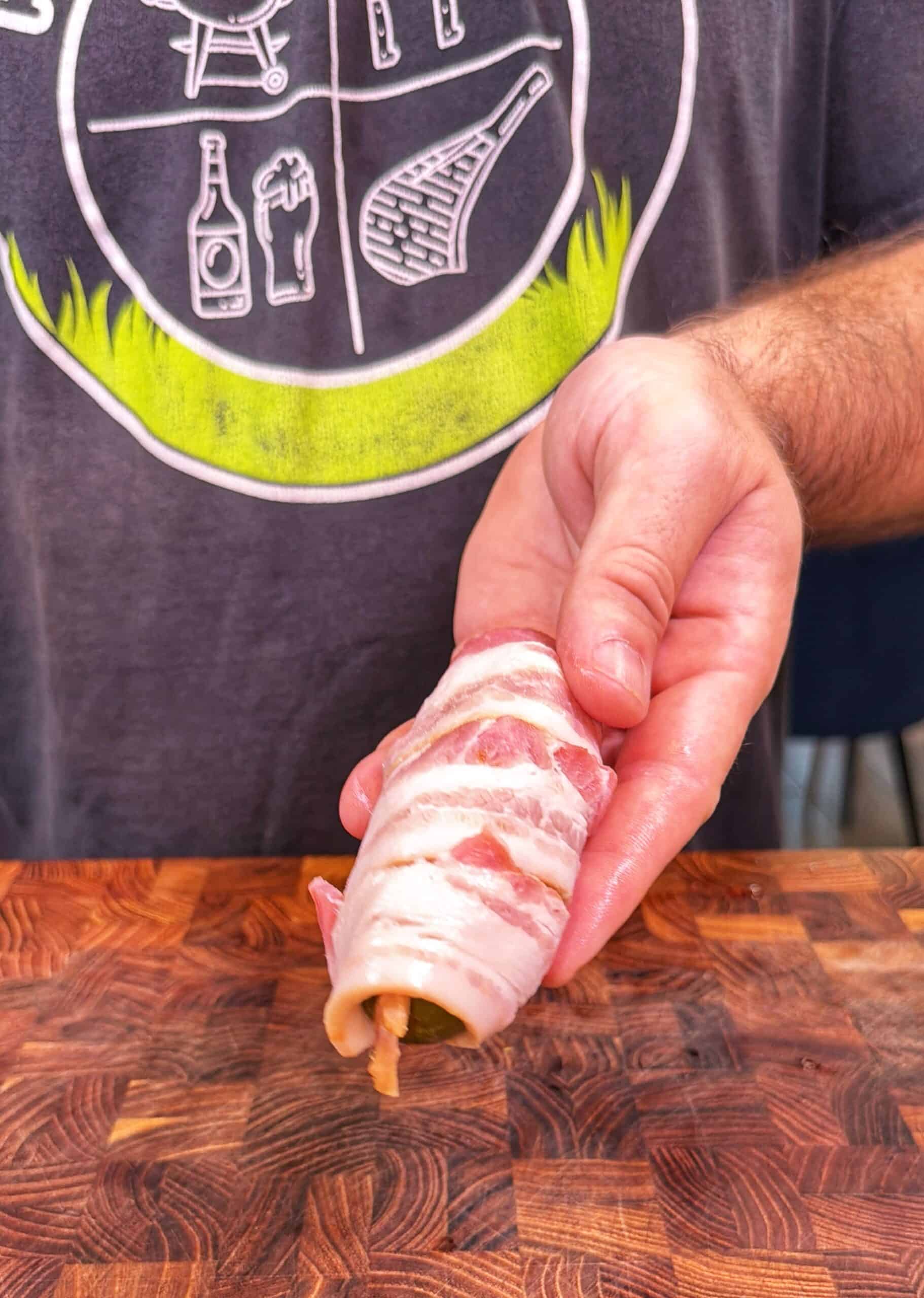 A hand holds a jalapeño stuffed with cheese and wrapped in bacon, ready to cook, over a wooden <a href=