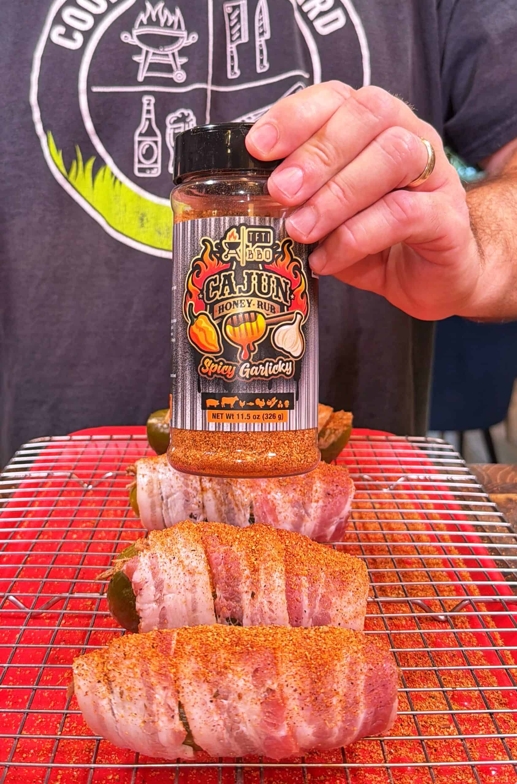 A hand holds a jar of Cajun honey rub over bacon-wrapped jalapeños on a wire rack, ready for grilling.