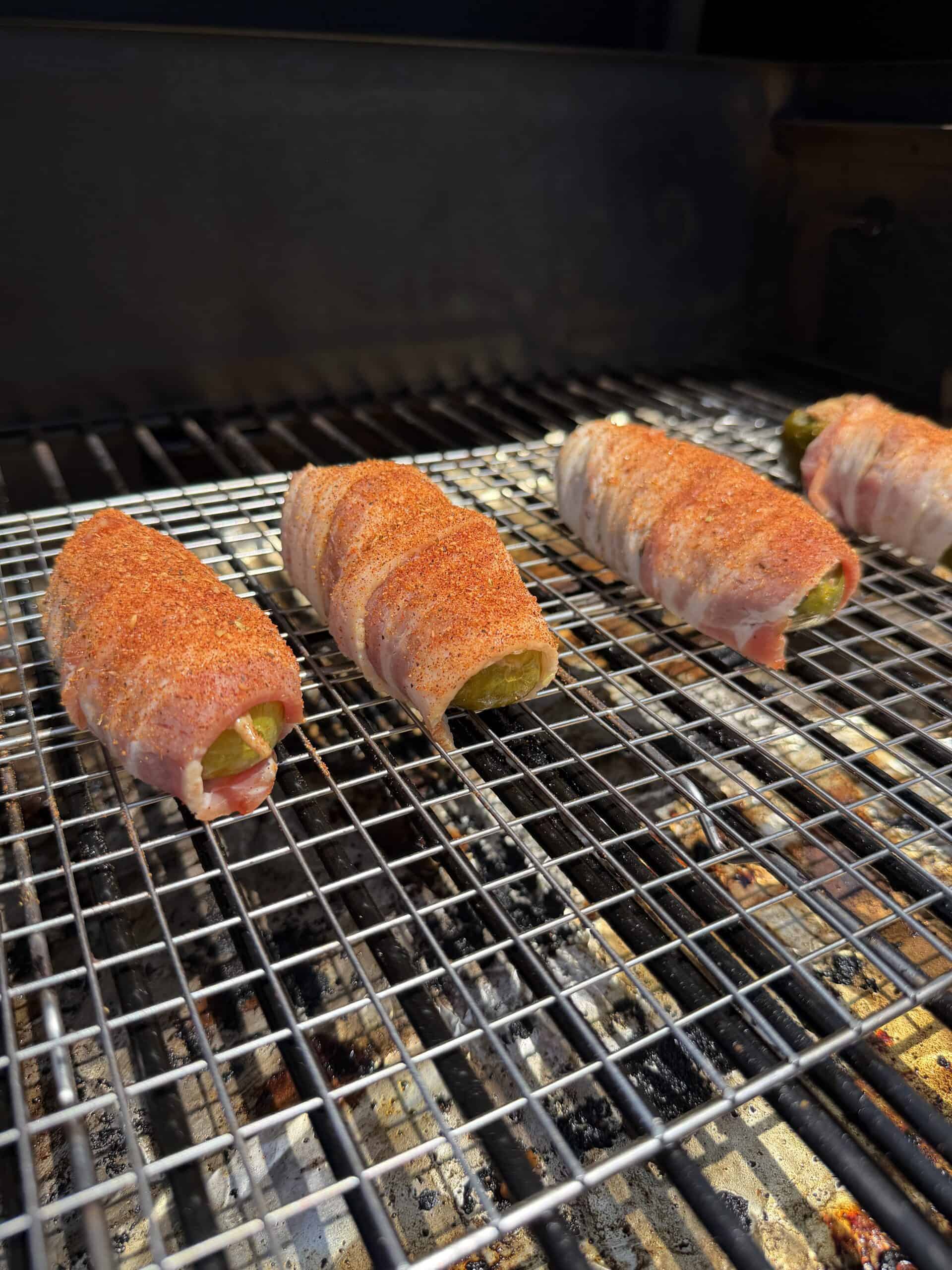 Four bacon-wrapped, spice-coated jalapeños are grilling on a wire rack, with the bacon crisping under the heat and the <a href=