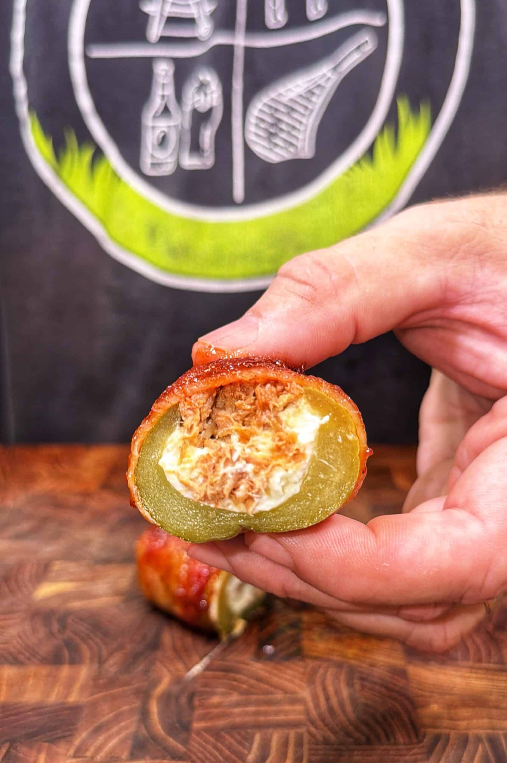 A hand holds a sliced jalapeño stuffed with shredded meat and cream cheese, wrapped in bacon. A wooden surface and a chalkboard drawing are in the background.