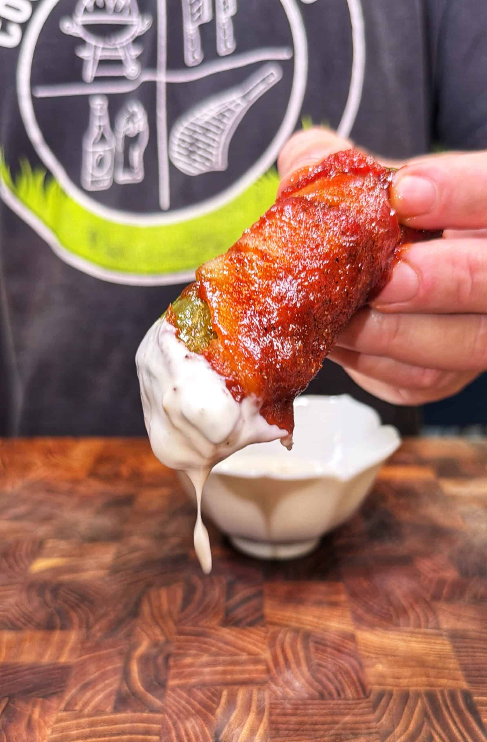 A hand holds a bacon-wrapped jalapeño popper dipped in creamy white sauce, with sauce dripping down. A wooden surface and part of a shirt with grilling graphics are visible in the background.