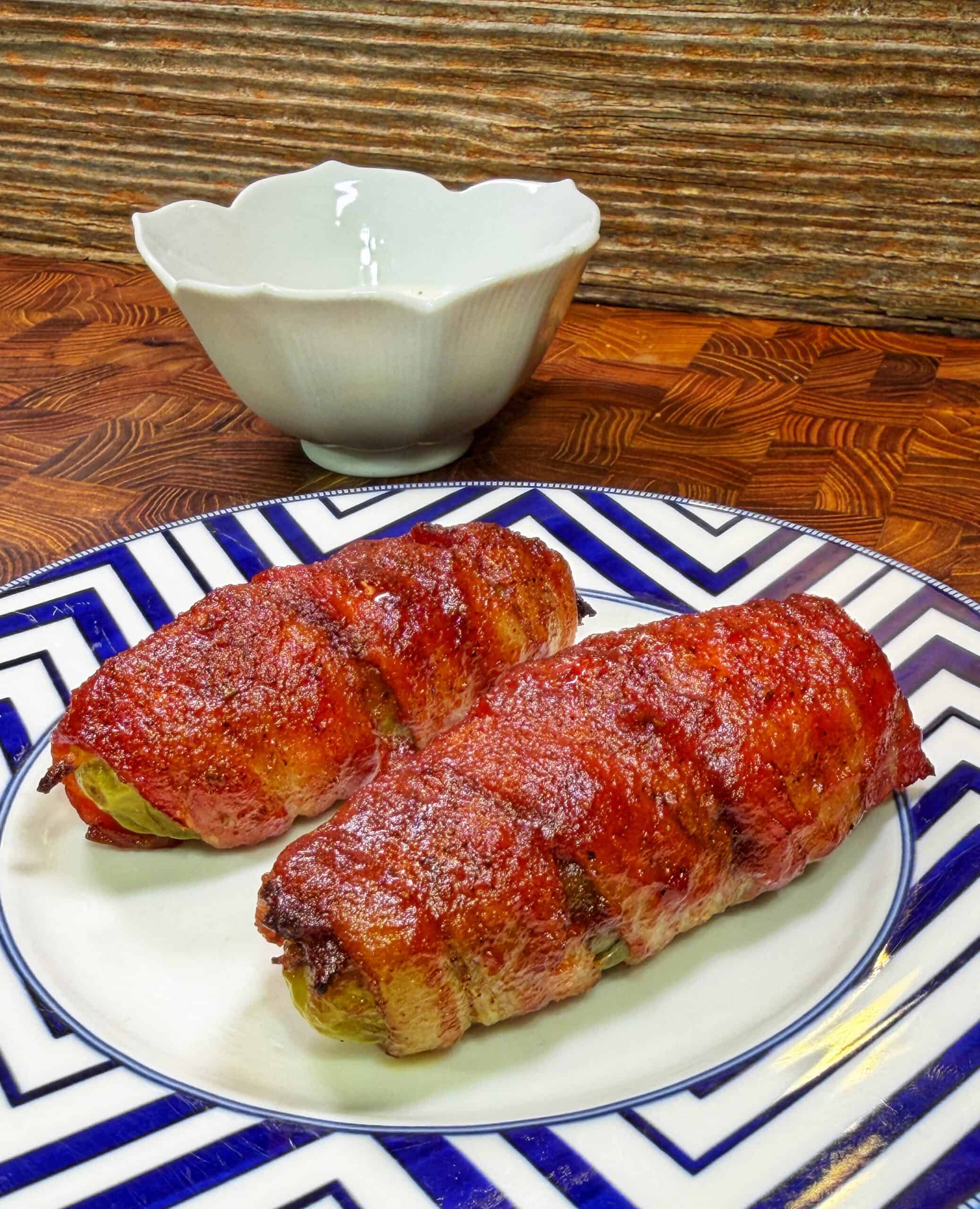 Two pieces of bacon-wrapped food sit on a patterned plate. A white bowl is in the background on a wooden surface with a ridged backdrop.