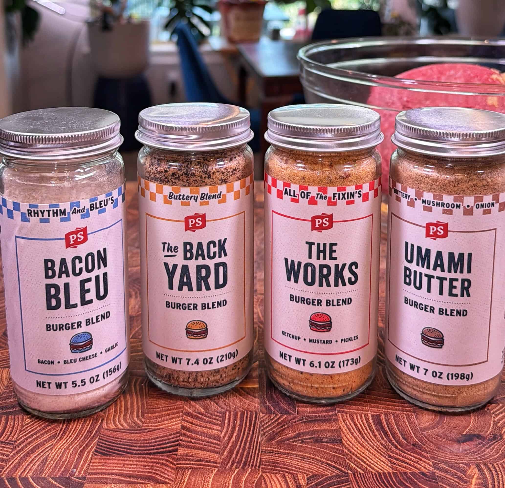 Four jars of PS burger blend seasonings—Bacon Bleu, The Back Yard, The Works, and Umami Butter—are lined up on a wooden surface with blurred kitchen items in the background.