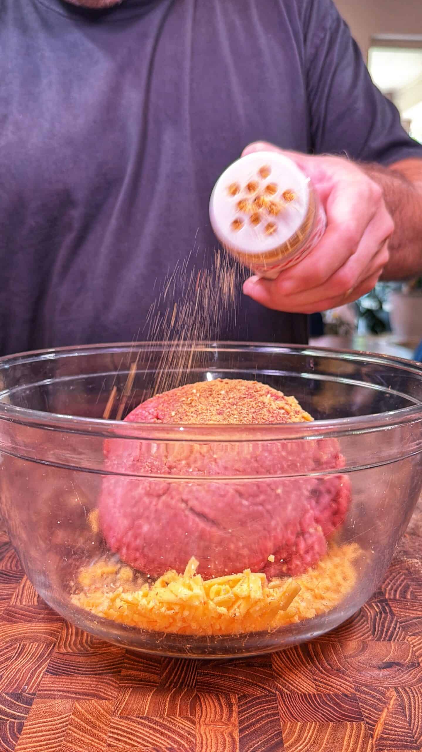 A person sprinkles seasoning onto raw ground meat in a glass bowl, which also contains shredded cheese, on a wooden surface.