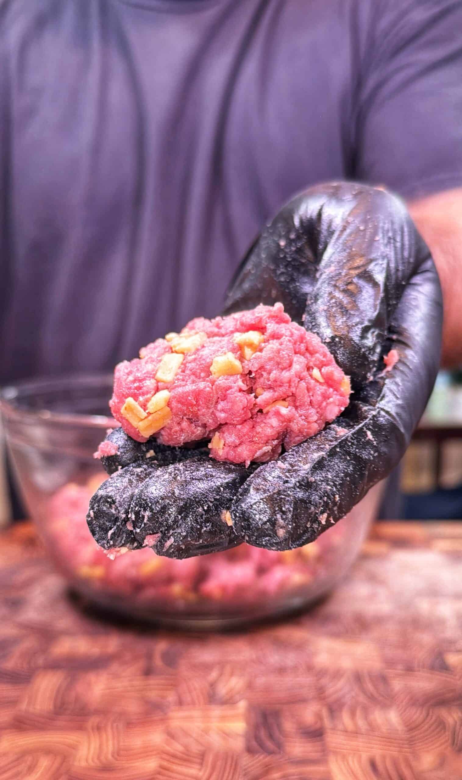 A person wearing a black glove holds raw ground meat mixed with chopped onions over a glass bowl, preparing food on a wooden surface.