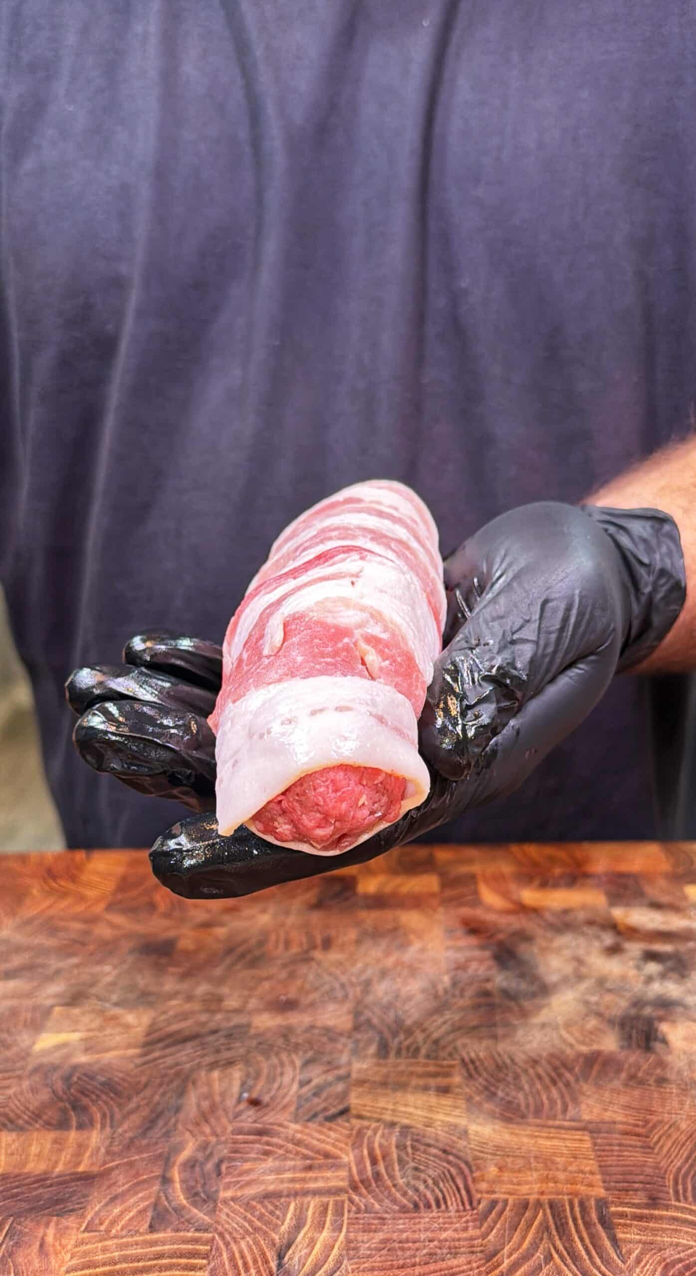 A person wearing black gloves holds a raw piece of meat wrapped in bacon over a wooden cutting board.