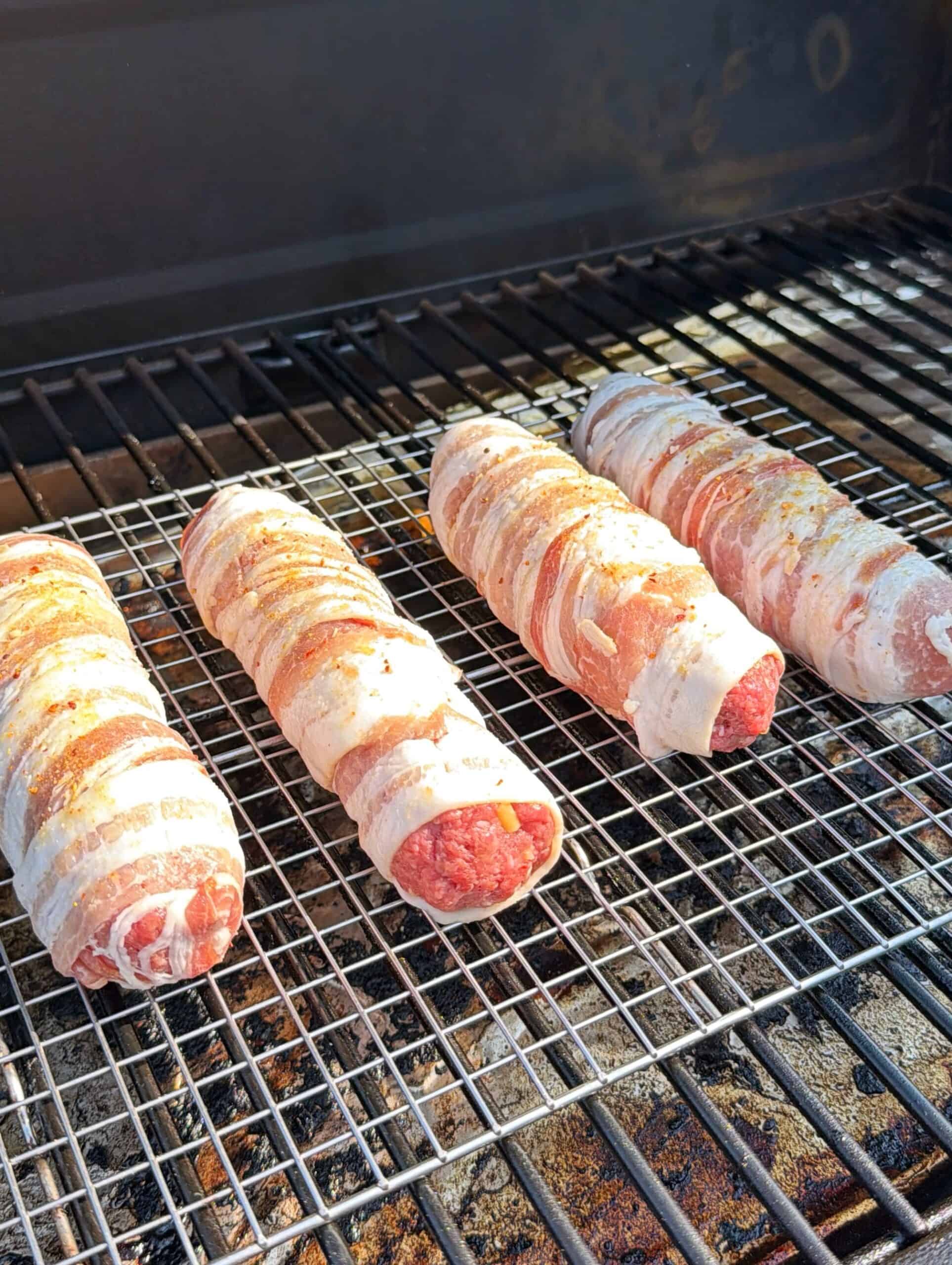 Four bacon-wrapped, sausage-shaped meat rolls are being grilled on a wire rack inside a barbecue grill, with sunlight highlighting their texture and seasoning.