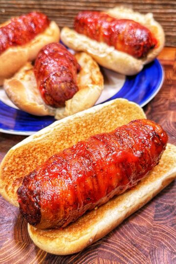 Four hot dogs wrapped in crispy bacon sit in toasted buns, with three on a blue-rimmed plate and one in the front on a wooden surface, all glazed with a shiny sauce.