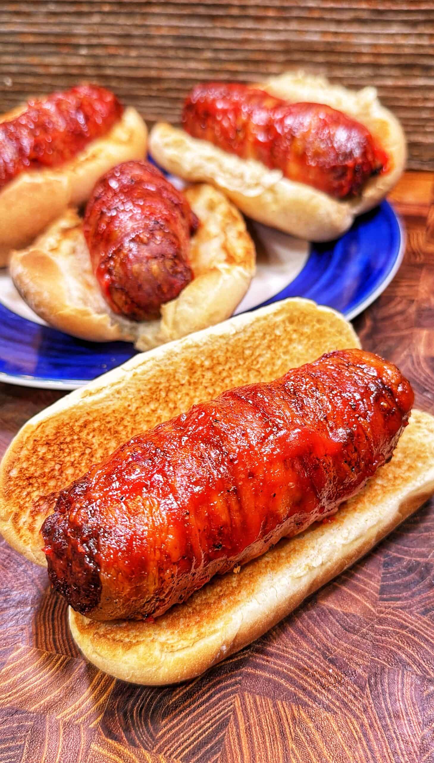 Four hot dogs wrapped in crispy bacon sit in toasted buns, with three on a blue-rimmed plate and one in the front on a wooden surface, all glazed with a shiny sauce.