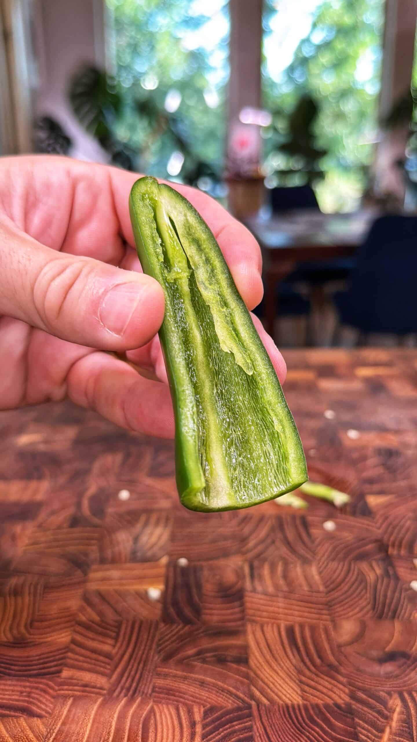 A hand holds a green jalapeño pepper sliced in half lengthwise over a wooden <a href=